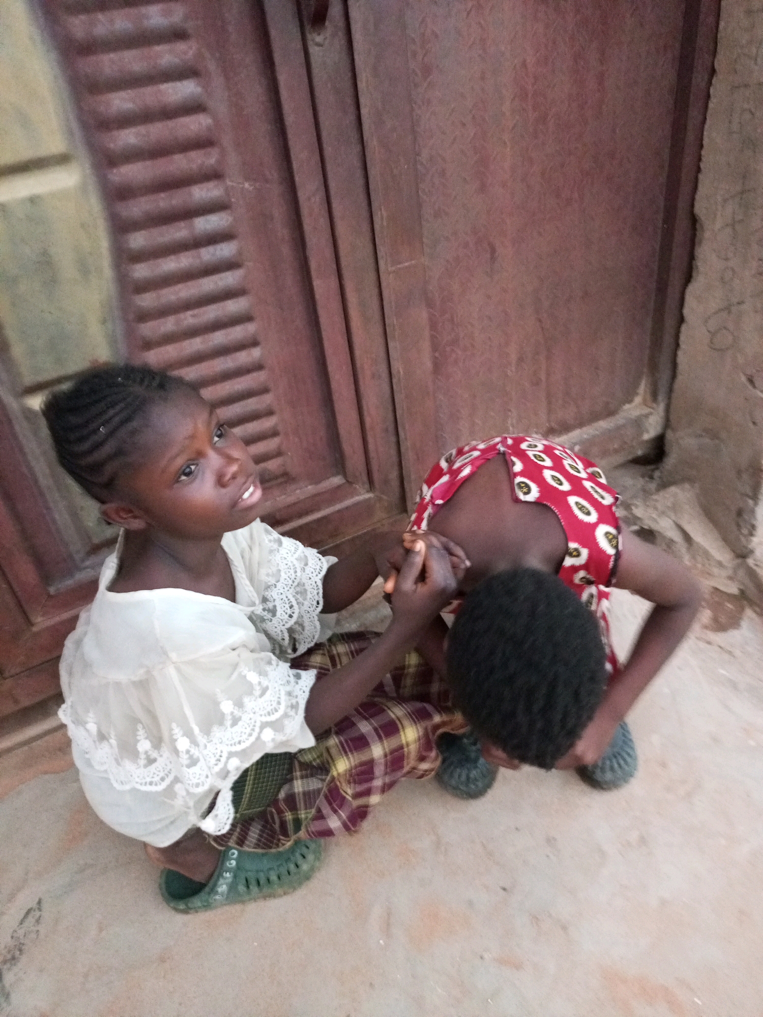 In Tempure, Nigeria, two young children are seen sitting on a dusty, uneven ground in front of a weathered, red wooden door. The child on the left, a girl, is looking upwards and slightly to the right, her face showing an expression of mild surprise or questioning. She has dark skin, braided hair, and is wearing a white lace-trimmed top and a plaid skirt. On her feet, she wears green sandals. The child on the right, whose face is mostly obscured, is bent over with their head down, appearing to be engaged with something on the ground or interacting with the other child. This child wears a red and white patterned garment. The environment suggests an outdoor setting with visible textures of the ground and the door.