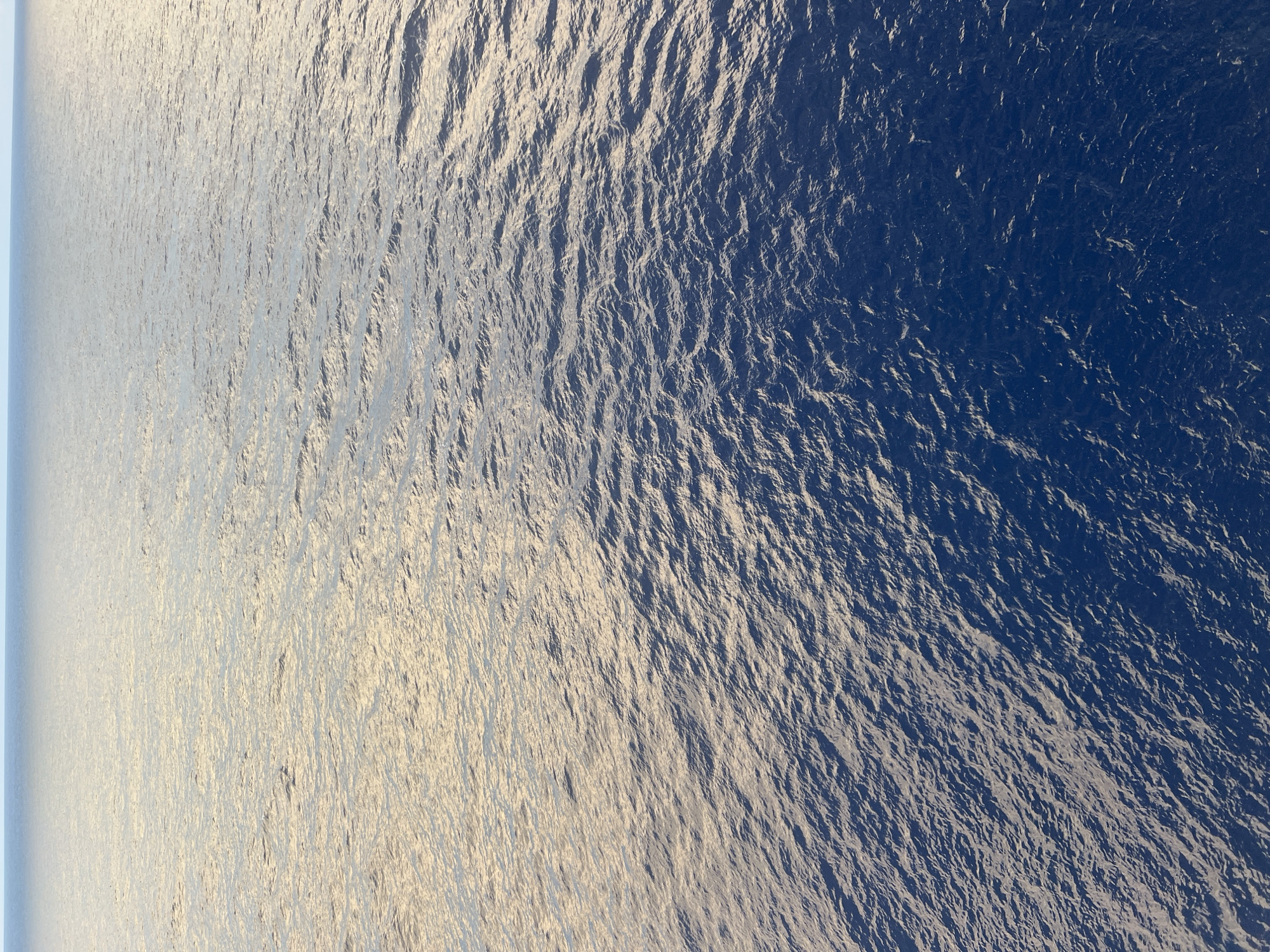 The image shows a vast expanse of calm ocean water under a bright sky. The water transitions from a lighter, golden hue on the left side of the frame to a deep blue on the right. The surface of the water is gently rippled, reflecting the light and creating a textured appearance. The horizon line is clearly visible, separating the water from the pale blue sky. There are no discernable objects or subjects in the water, and the scene is devoid of any human presence or activity. The overall impression is one of tranquility and openness. The location is Unknown, Unknown.