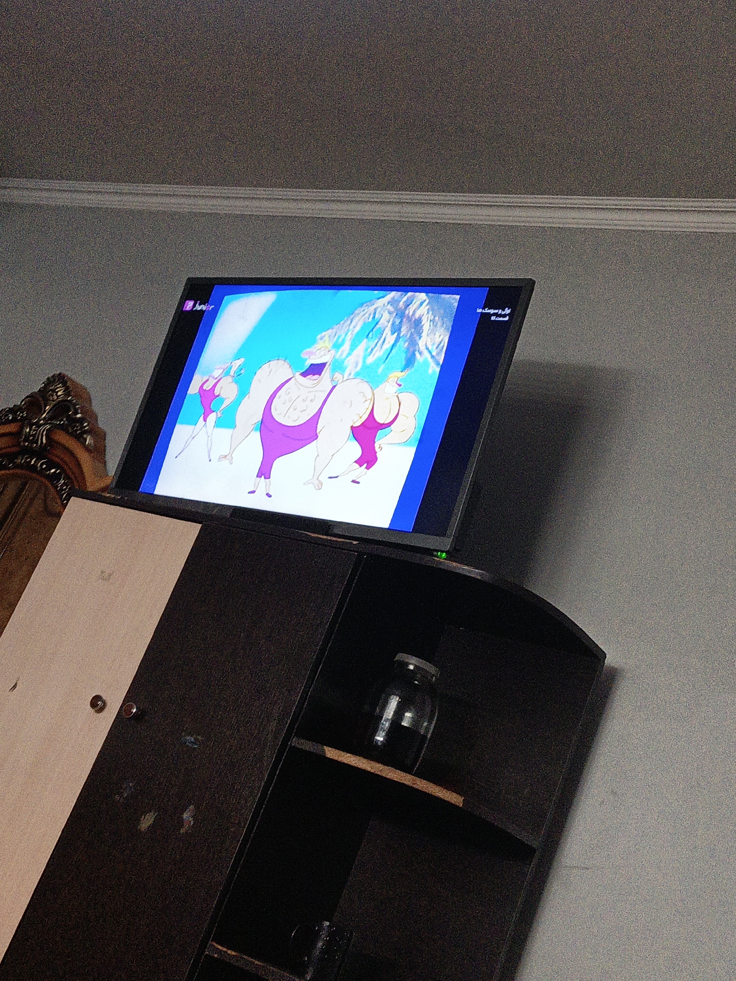 This is a medium-shot, eye-level, indoor, eye-level photograph taken from a low angle, showcasing a television screen displaying an animated cartoon with muscular characters in purple swimsuits. To the left of the television is a portion of a wooden cabinet with a carved wooden headboard. Below the television is a dark wooden cabinet with a shelf containing a glass jar. The overall scene is dimly lit, with a wall painted a neutral gray. The television is tilted forward and shows content from a channel called "Junior". The scene is located in Pistamazor, Tajikistan.
