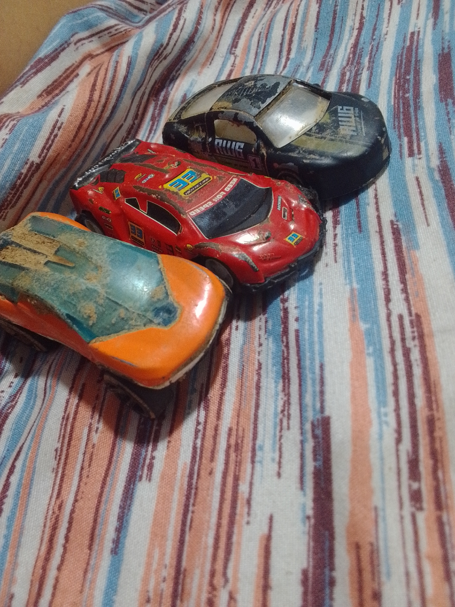Three toy cars are arranged on a striped fabric surface. The cars are dusty and appear to be worn. The fabric has vertical stripes of white, light blue, peach, and dark red. The image is taken from a slightly elevated perspective, looking down at the cars. There are no people or other discernible objects in the scene. The location in La Tagua, Venezuela is not indicated by any visual cues in the image.
