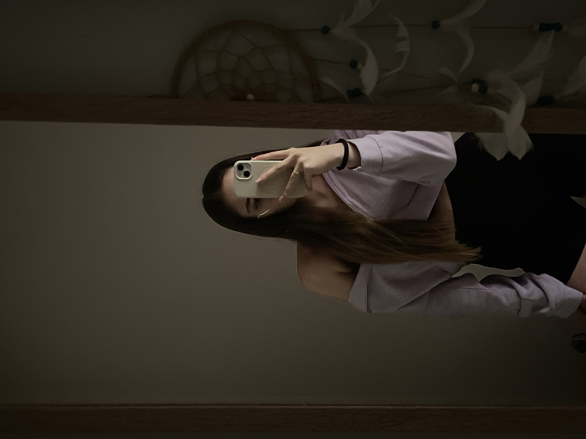 The image shows a person taking a mirror selfie. The person is holding a white smartphone, obscuring their face. They are wearing a light purple, off-the-shoulder sweatshirt and black shorts. The mirror reflects a wall with a dreamcatcher hanging above the mirror. The location appears to be indoors, in a dimly lit room. The location is Unknown, Unknown.