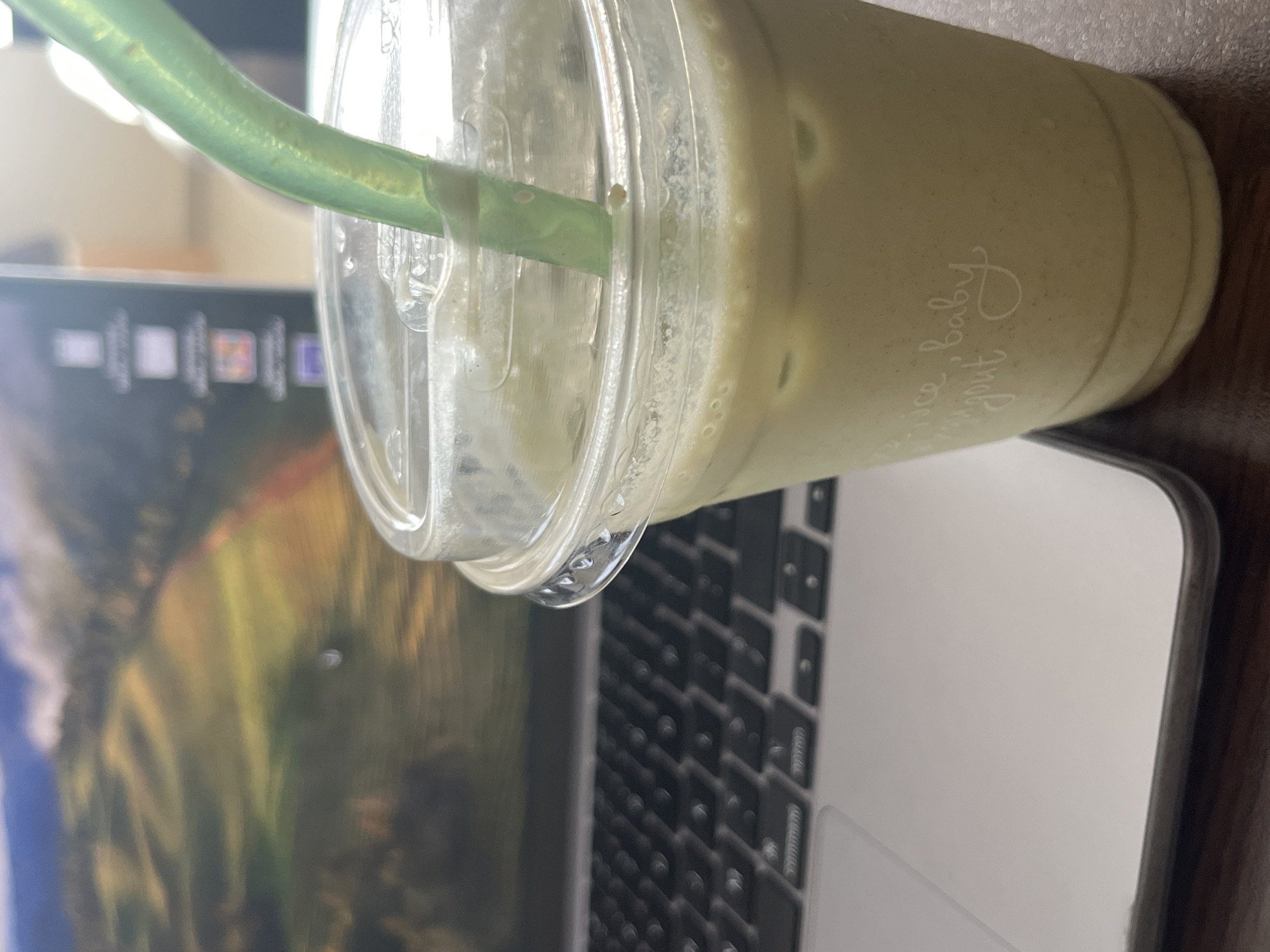 A close-up photograph shows a partially consumed beverage in a clear plastic cup with a lid and a green straw, resting on a laptop computer. The beverage appears to be a light-colored smoothie or shake. The laptop screen displays a blurred image of a landscape. The photo appears to be taken indoors on a desk or table with a brown surface. The text on the cup reads "...juice baby... yogurt". The location in Sofia, Bulgaria, is not apparent from the image.