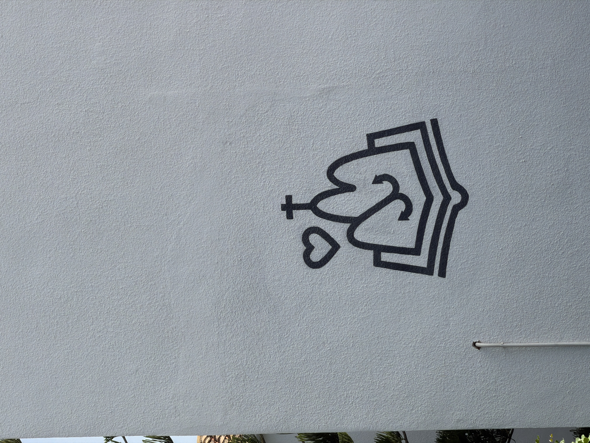 This is a medium shot, eye-level, outdoor, natural light photograph of a black line drawing on a light grey, textured wall. The drawing depicts an open book with two abstract, mountain-like shapes rising from its pages. A Christian cross sits atop the rightmost mountain, and a heart shape is to the left of the leftmost mountain. A white pipe runs vertically up the wall to the left of the drawing. In the far left background, the top of a building with terracotta tiles and some green foliage are visible.