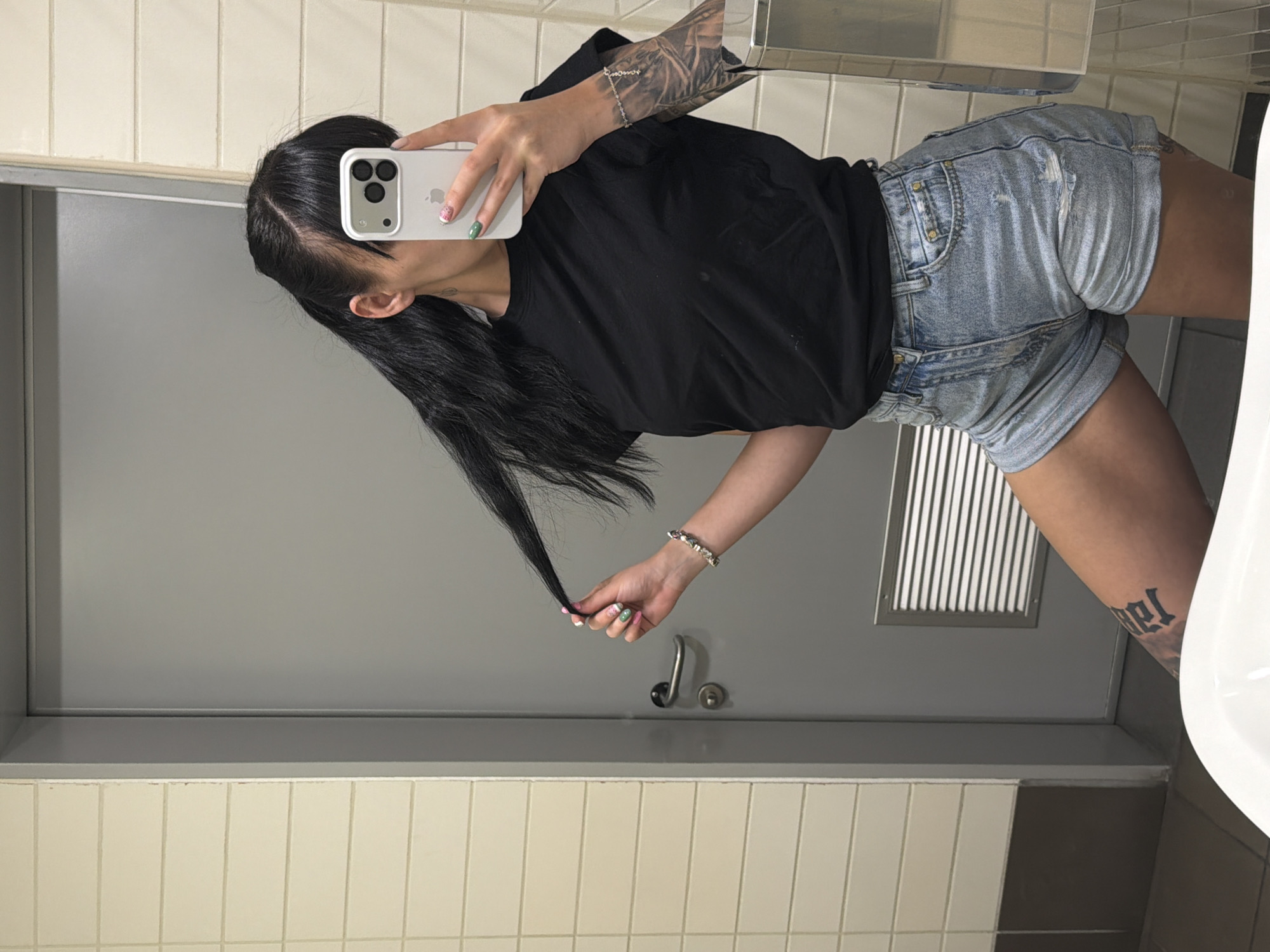 A person with long, dark hair is captured in a mirror selfie within what appears to be a bathroom or public restroom. The individual wears a black short-sleeved t-shirt and light blue distressed denim shorts. A white iPhone, held in their right hand, obscures their face, revealing its multi-lens camera module and an Apple logo visible through a case cutout.
Their right forearm features an intricate, full-sleeve tattoo. On their left wrist, a silver-toned bracelet with small charms is visible. The person's left hand holds a strand of their hair, showcasing manicured nails with distinct designs, including a small flower on one and a green pattern on another. A dark, gothic-style script tattoo is partially visible on their left thigh.
The setting consists of light beige square tiles on the lower walls, transitioning to a plain grey surface above. Reflected in the mirror are a grey door, a metal vent or grille on the lower wall, and a white ceramic fixture (likely a toilet or urinal) to the right. A curved metal handle is also seen on the grey wall. The lighting suggests an indoor environment.