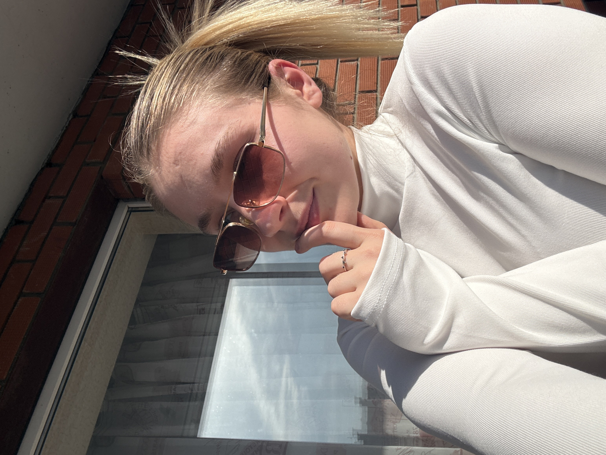 A person with fair skin and blonde hair pulled back in a ponytail is wearing sunglasses with a gold frame and brown lenses and a white turtleneck top. They are resting their chin on their finger, which has a ring on it. The background shows a brick wall, a window with blinds, and a sunny sky. It is difficult to provide the location in Izhevsk, Russia without more information about the surroundings or landmarks visible in the image.