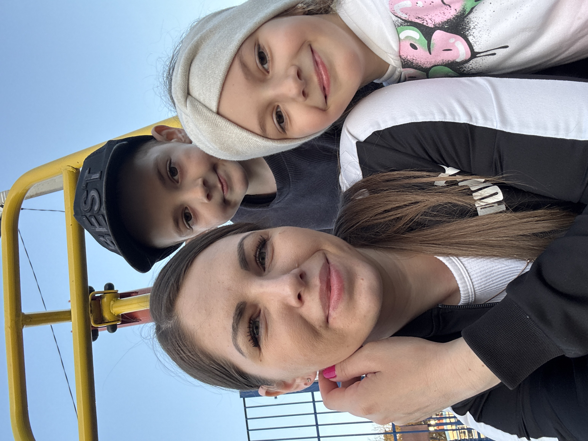 A selfie taken from a low angle of a woman with brown hair and pink nail polish, a young boy in a black baseball cap, and a young girl wearing a white headband. The woman is wearing a white ribbed top and a black and white track jacket. The girl is wearing a white sweatshirt with a colorful graphic. A yellow metal structure is visible in the background. The location is in Раздолье, Russia.