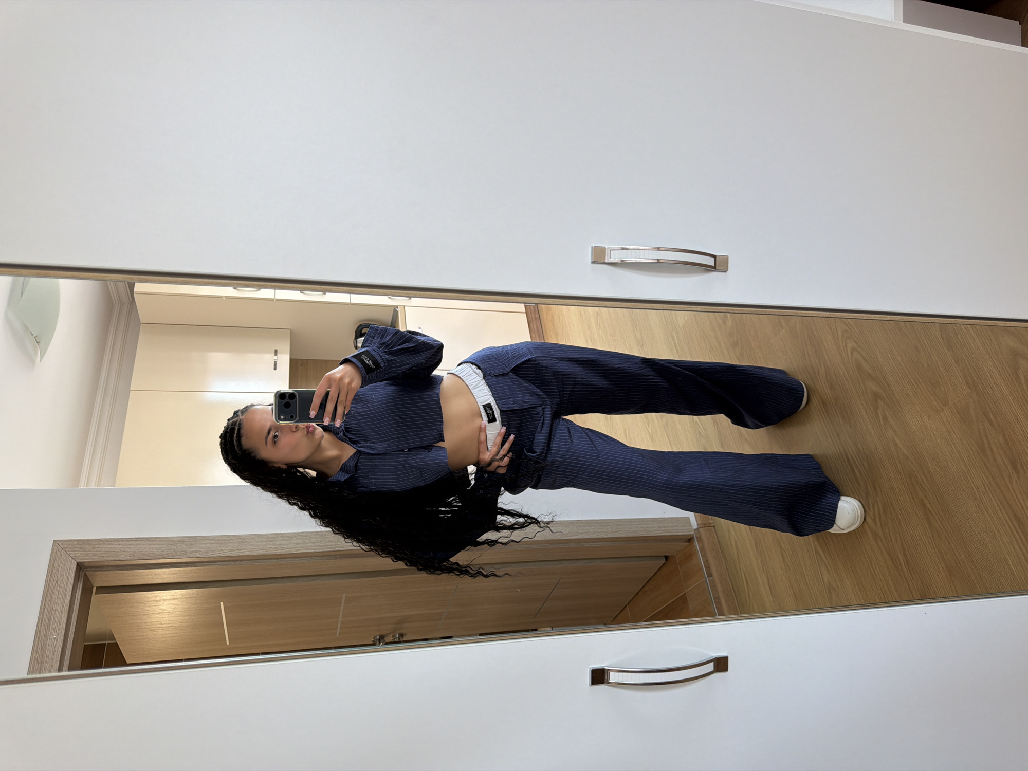 A young woman with long, dark, braided hair is standing in a room, taking a selfie in a full-length mirror. She is wearing a navy blue pinstripe crop top and matching wide-leg trousers with a white elastic waistband. She has her left hand on her hip and her right hand holding a smartphone. The room appears to be a bedroom or dressing area, with a white wardrobe to the right, a wooden door on the left, and light-colored kitchen cabinets visible in the mirror's reflection. The floor is made of wood laminate. The scene is indoors and appears to be in an apartment. It is not possible to determine the city of Nesebar, Bulgaria from the visual information provided.