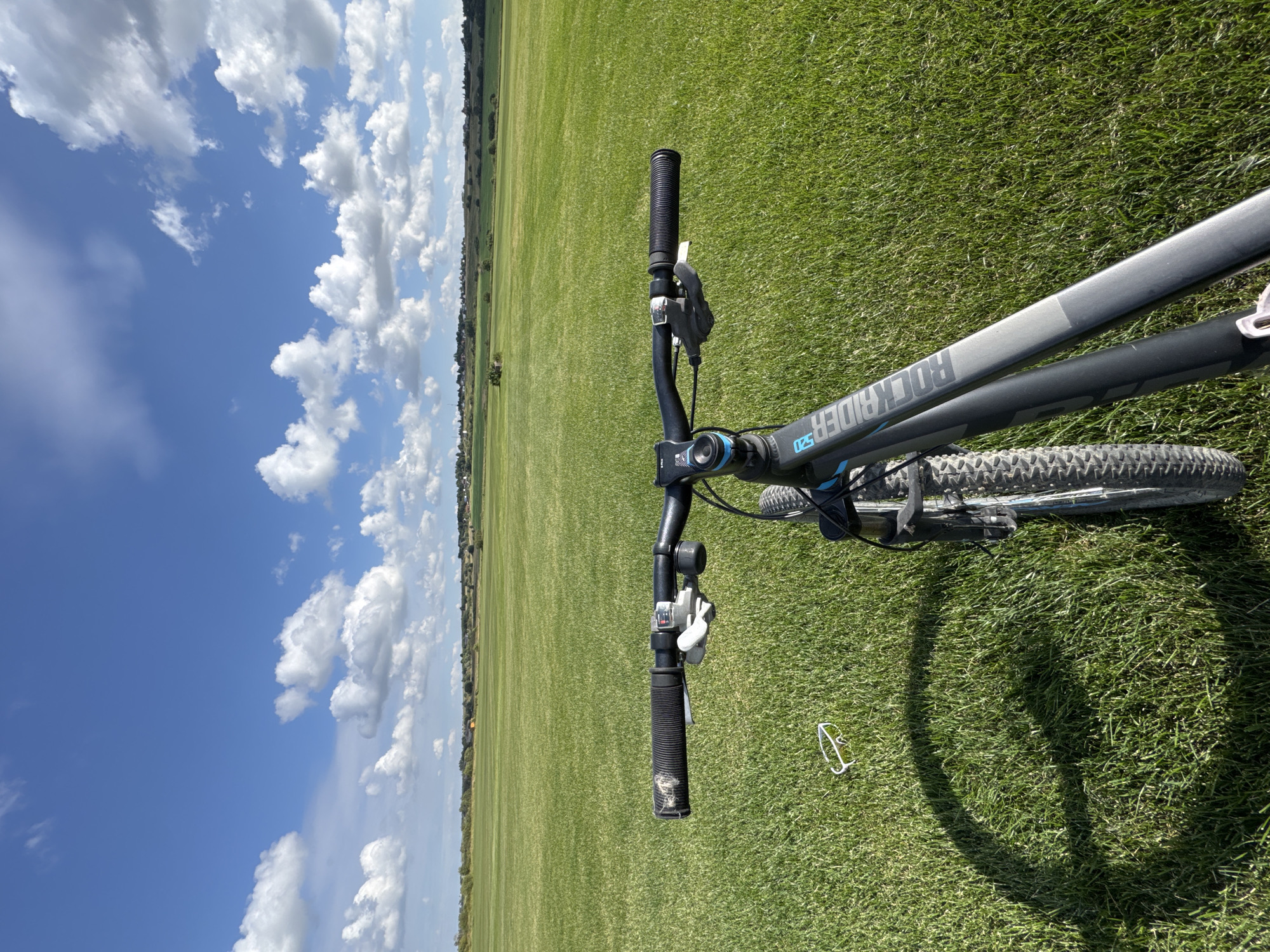 A first-person perspective shot captures the handlebars and front of a mountain bike parked on a vast, flat, green field under a bright blue sky dotted with fluffy white clouds. The bicycle is dark gray with blue accents, and the word "ROCKRIDER" is visible on the frame. A pair of white sunglasses lies on the grass to the left of the bike, casting a shadow. In the distance, a line of trees and small houses marks the horizon. The scene appears to be in or near Kempelevo, Russia, given the expansive agricultural landscape and typical Russian architecture visible on the horizon.