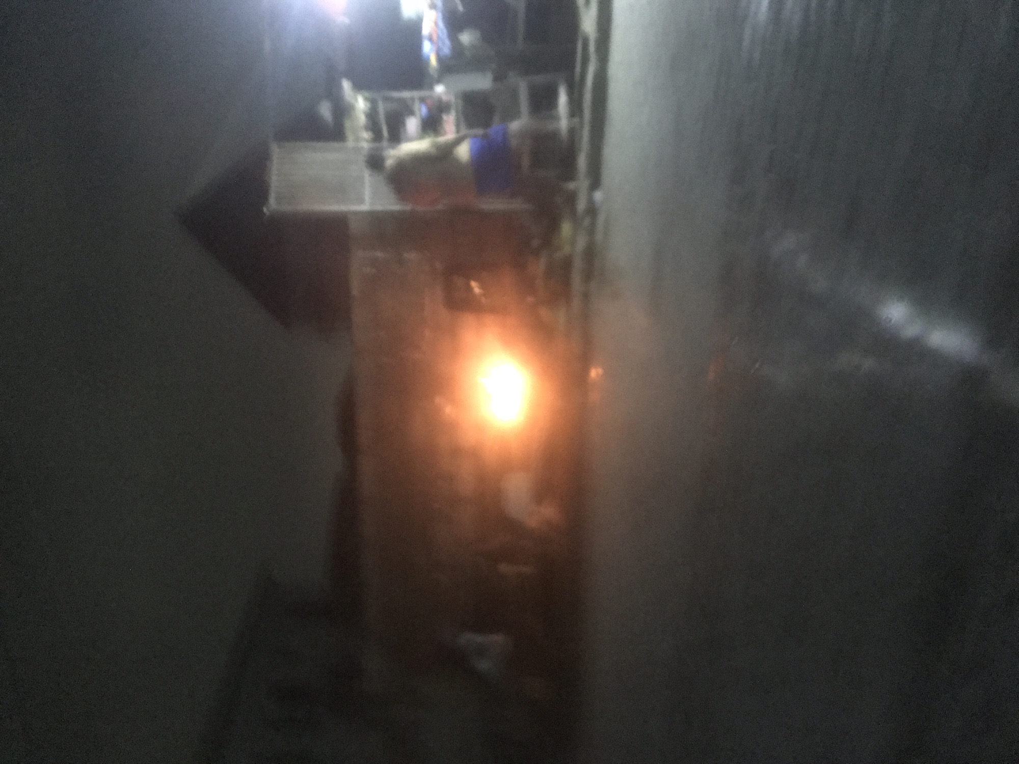The image depicts a street scene at night, likely in an urban environment. A bright, orange light source, possibly a fire or a powerful lamp, illuminates the scene from the left side. Several blurry figures are visible in the background. To the right, a person wearing a dark top and blue lower garment is standing near a structure that appears to be a stall or market stand, with shelves stocked with items. A large, dark, triangular shape, resembling a roof or a building, looms in the background. The ground is wet and reflective, suggesting recent rain or damp conditions. The overall atmosphere is dark and somewhat hazy, with artificial lighting creating strong contrasts.