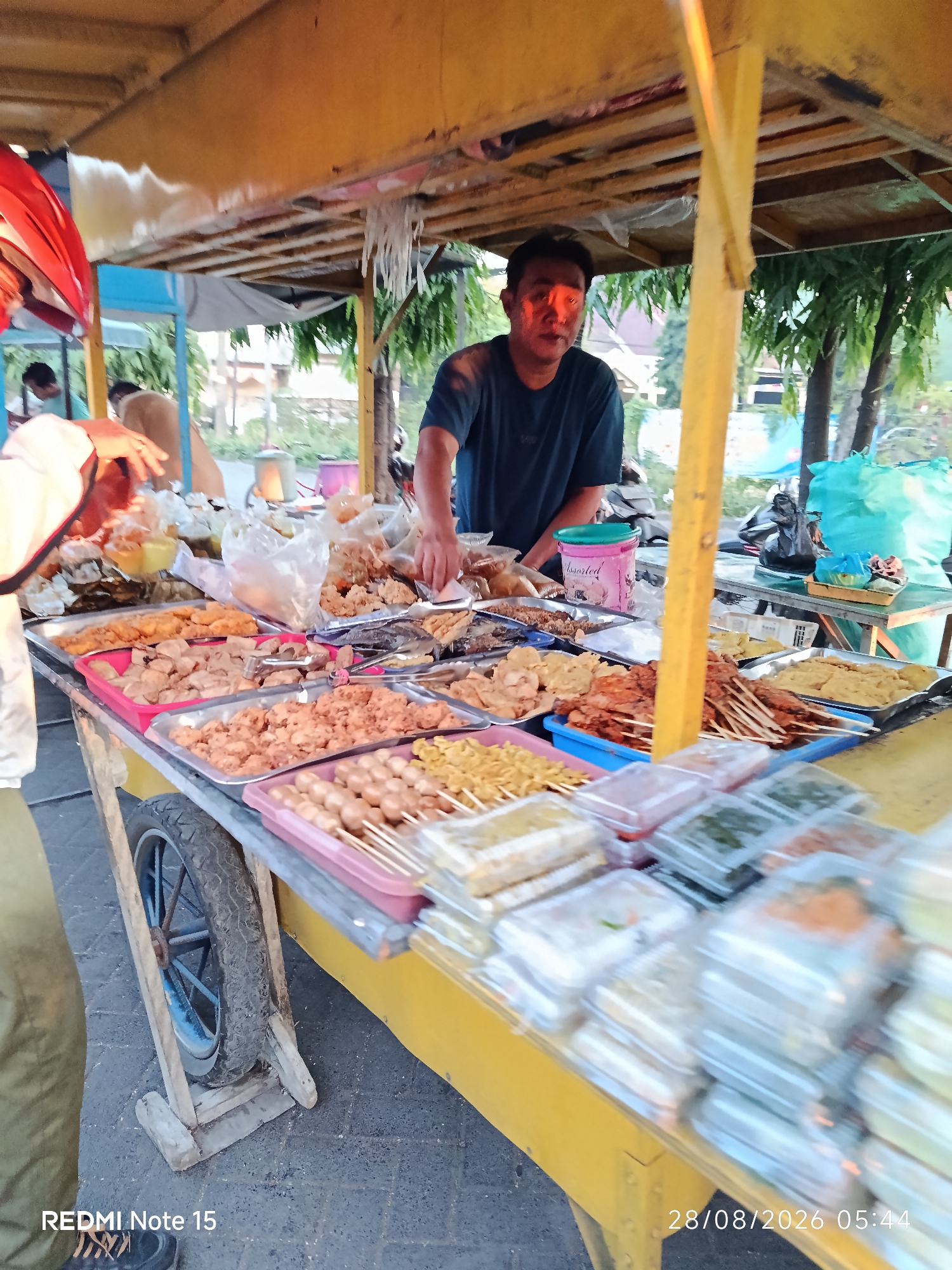 The image depicts a street food vendor stall in Keboansikep, Indonesia. A man, likely the vendor, is seen from the chest up, wearing a dark t-shirt, with his face illuminated by a bright light source, possibly a street lamp or the setting sun. He is standing behind a mobile food cart laden with various fried snacks displayed in trays and plastic containers. These snacks include what appear to be fried chicken pieces, meatballs on skewers, and other deep-fried items. The cart itself is constructed with yellow painted metal and wood, with a large wheel visible on the left side. The background suggests an outdoor setting, with trees and foliage visible, indicating a potentially urban or semi-urban environment. Other blurred figures and a motorcycle are partially visible in the background. The overall scene captures a moment of commerce and daily life in a local Indonesian setting.