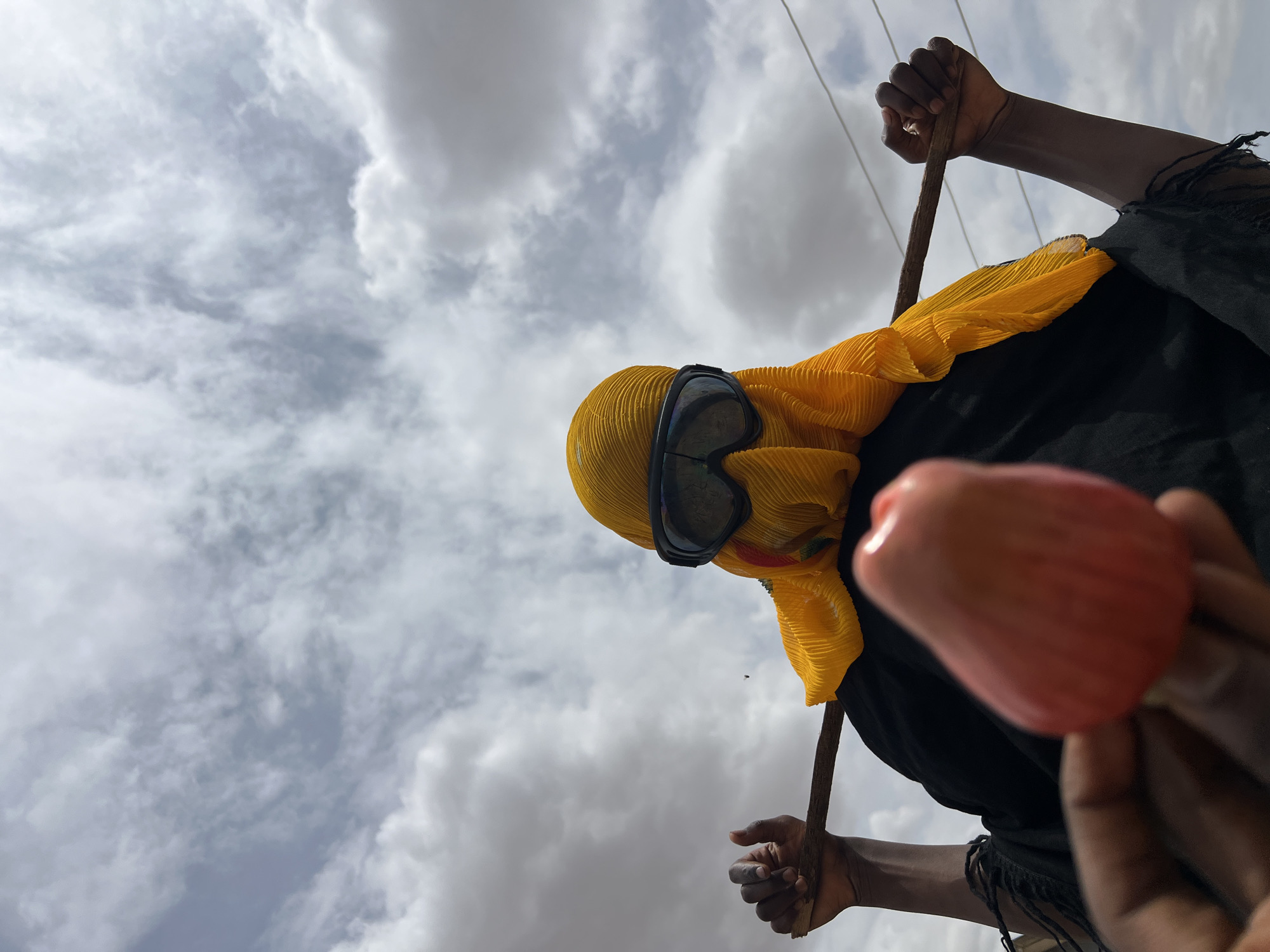 A low-angle shot reveals a person dressed in black attire with a vibrant yellow head covering, featuring a pair of goggles resting on their forehead. The person holds a brown stick in one hand and a reddish fruit in the other, against a backdrop of a cloudy sky. Thin electrical wires are visible against the sky. While the specific location within Udu, Nigeria is not determinable from the image, the scene suggests an outdoor setting with infrastructure present.