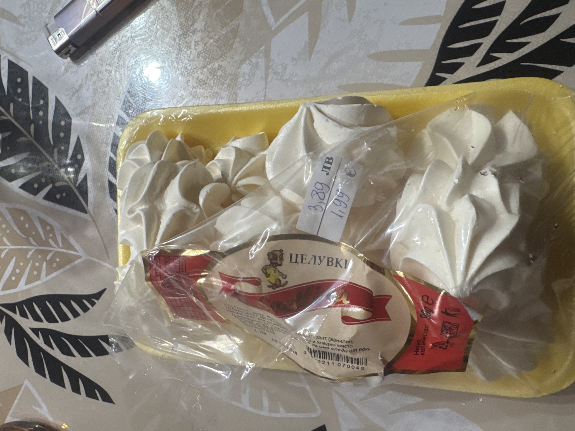 The image shows a yellow plastic tray of meringue kisses, commonly known as "Tseluvki" (целувки) in Bulgarian, wrapped in clear plastic. The product label is visible, featuring a teddy bear illustration and the product name in Cyrillic script. It also indicates the price, 3.89 Bulgarian Leva or 1.99 Euro. The label states that the product contains vanilla flavoring and should be stored in a cool, dry place. It also mentions that traces of nuts may be present. The net weight is listed as 90g. The background is a table with a patterned surface. A lighter is also visible on the table. Based on the product labeling in Bulgarian, it is plausible that this image was taken somewhere in Bulgaria. It is not possible to determine the specific city of Sarnevo, Bulgaria, from this image.