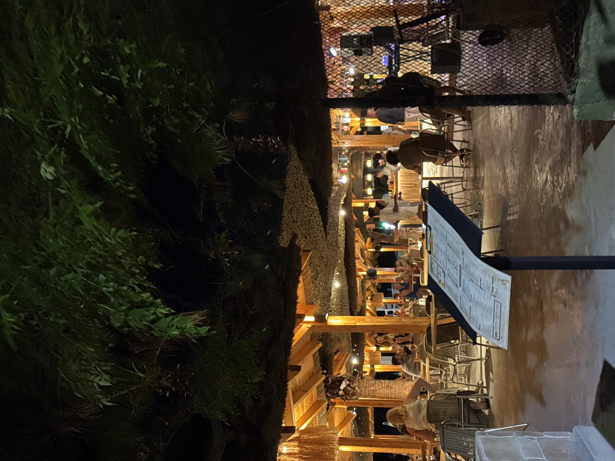 The image shows an outdoor dining area at night in Boreti, Montenegro. A thatched-roof structure covers multiple tables and chairs where patrons are seated. In the background, a bar area is visible with people standing and being served. String lights are visible under the roof, providing illumination. A menu board is placed in the foreground. The area appears to be a restaurant or beach club, with palm trees and foliage overhead.