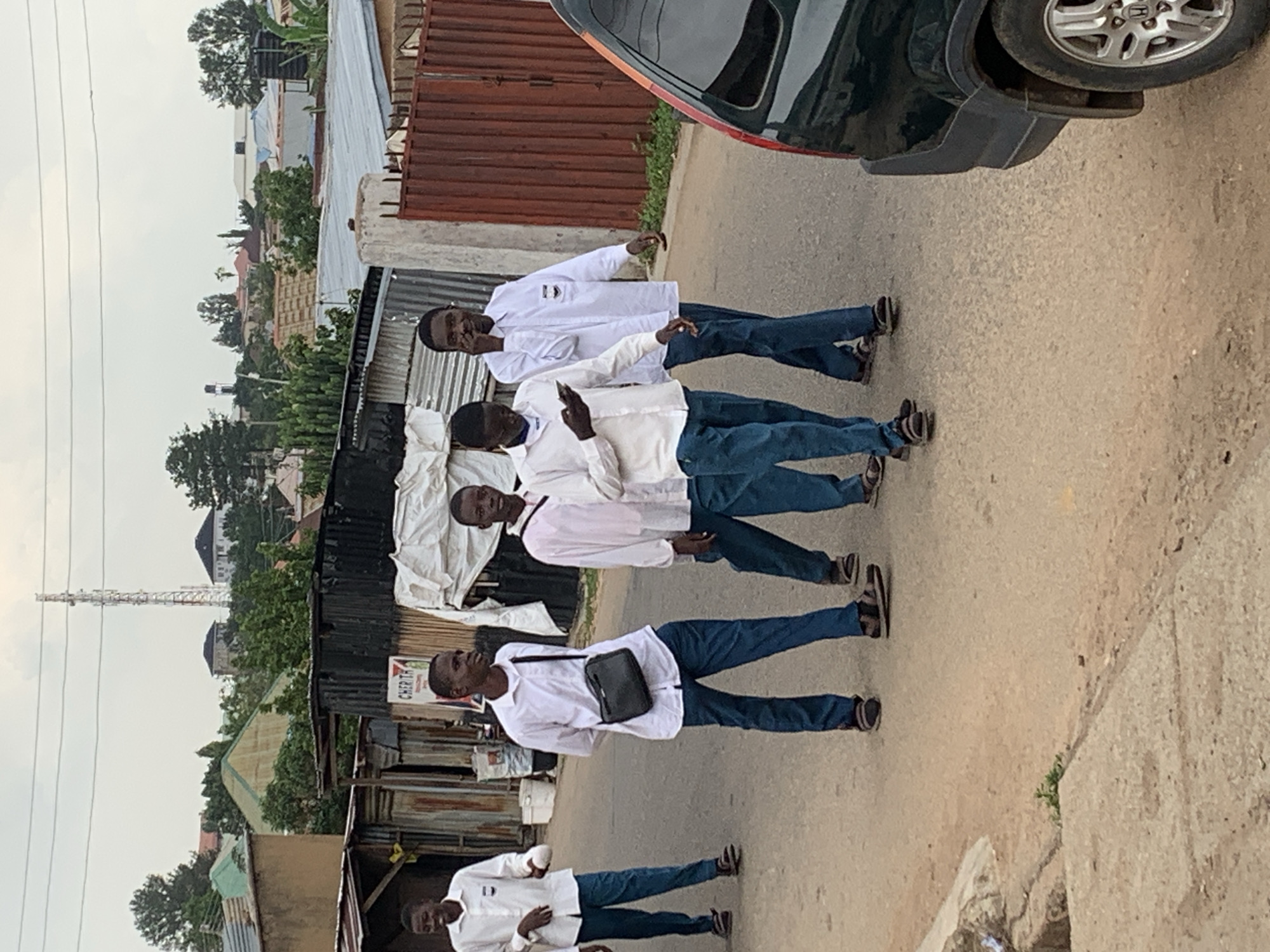 This image captures a group of five young men, likely students in school uniform, walking down a dusty, paved street. The young men are dressed in white shirts and blue trousers. The scene is set in Jos, Nigeria, as indicated by the presence of tin-roofed buildings, some constructed from corrugated metal, and a distinct cellular tower visible in the background, a common feature in Nigerian urban landscapes. The environment is typical of a residential area in Jos, with modest houses, some appearing to be under construction or in a state of disrepair. The men are walking in the middle of the street, with a dark SUV partially visible on the right side of the frame, suggesting a typical street scene in the city. The sky is overcast, indicating a potentially humid or rainy day. One of the young men in the center is carrying a black satchel across his body, and another on the right appears to be holding a mobile phone to his ear, possibly engaged in a conversation. The overall impression is one of everyday life unfolding in a Nigerian city.