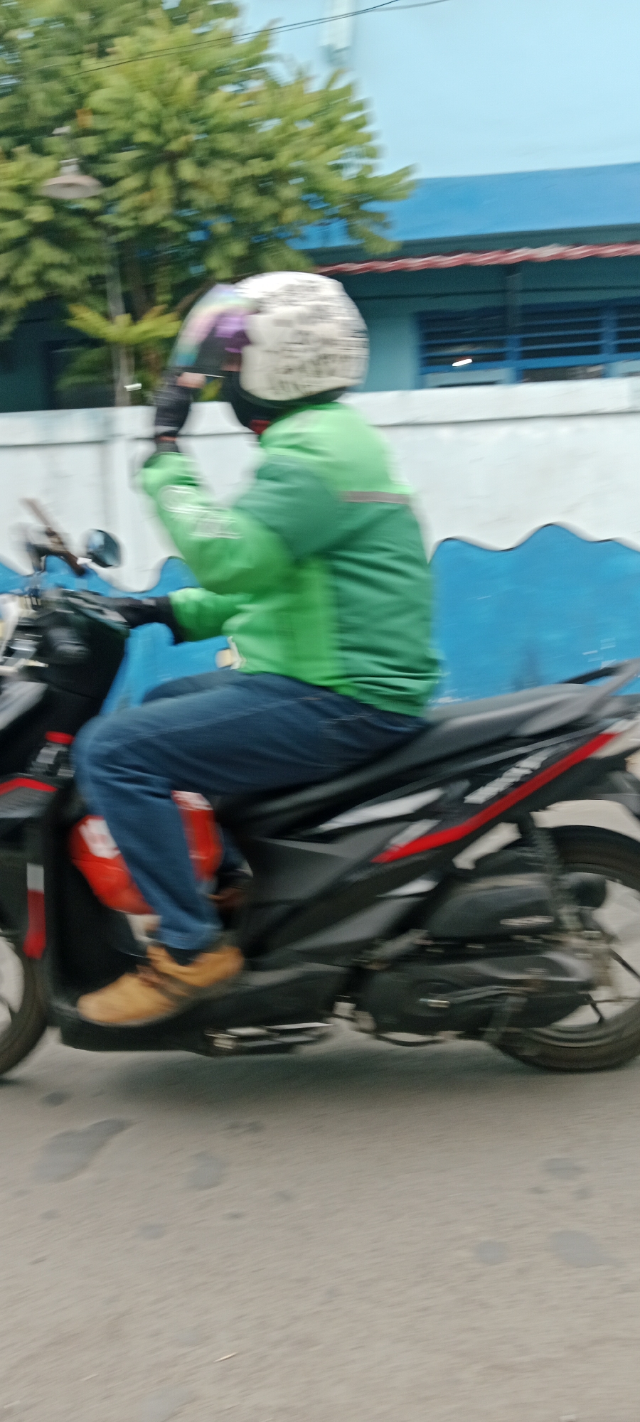 A person wearing a green jacket and a white helmet with a colorful design is riding a black motorcycle. The person is also wearing blue jeans and brown shoes. The motorcycle has red accents on its side. The background shows a blue building with a white wave-like pattern on its lower section and a tree with green leaves. The scene appears to be in the city of Purwomartani, Indonesia, as indicated by the presence of a street with some water spots and a light pole in the distance. The image is slightly blurry, suggesting motion.