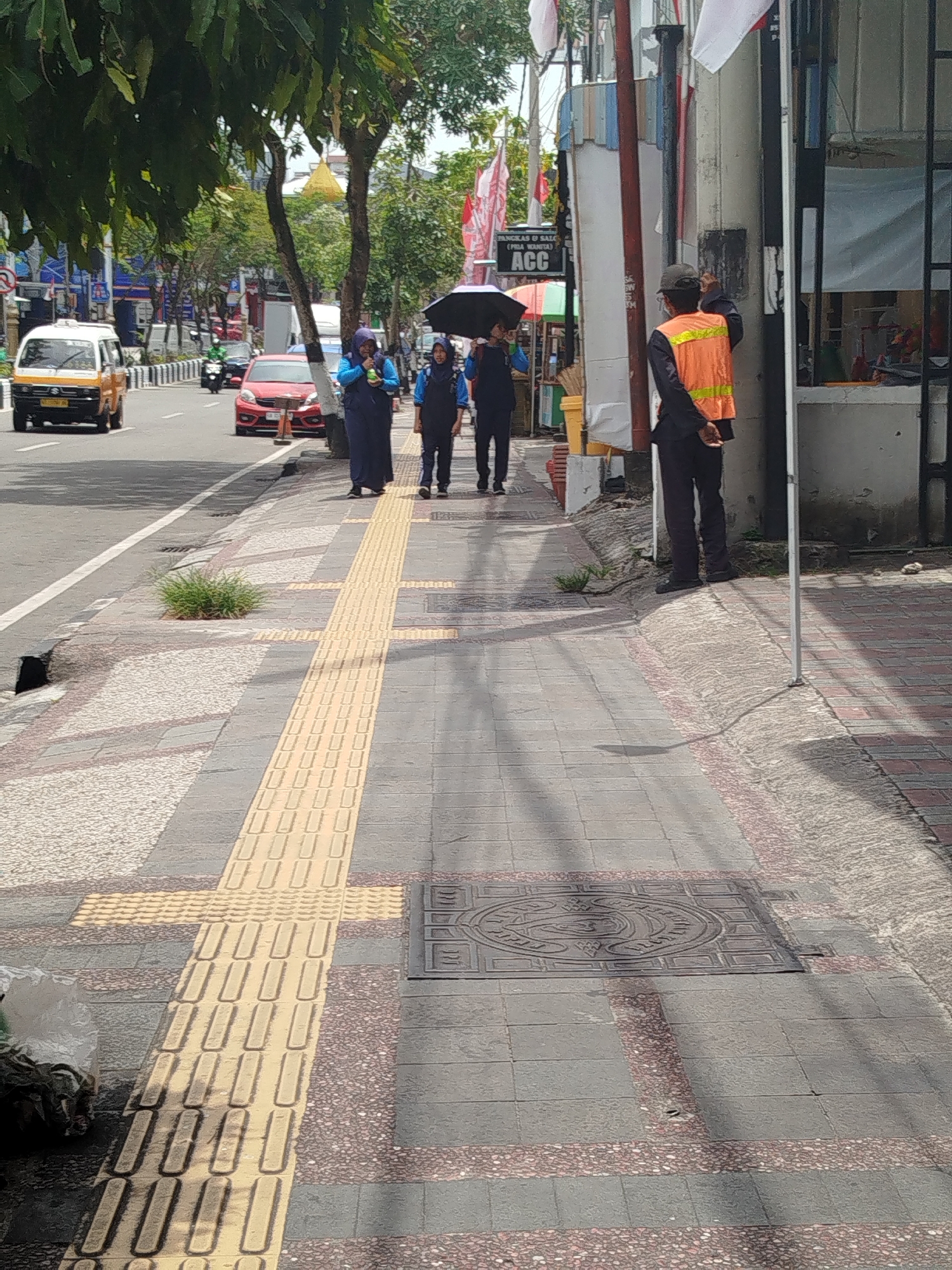 The image depicts a sidewalk scene on a sunny day in Balikpapan, Indonesia. A man in an orange vest and dark pants stands with his back to the camera, appearing to be observing the street. In the background, three individuals walk along the sidewalk, two of whom are women wearing hijabs. One woman is holding a black umbrella. The sidewalk is paved with textured tiles, including tactile paving strips for the visually impaired. A street with traffic, including a yellow and white vehicle and a red car, is visible to the left. Trees and buildings line the street. A manhole cover is present on the sidewalk.