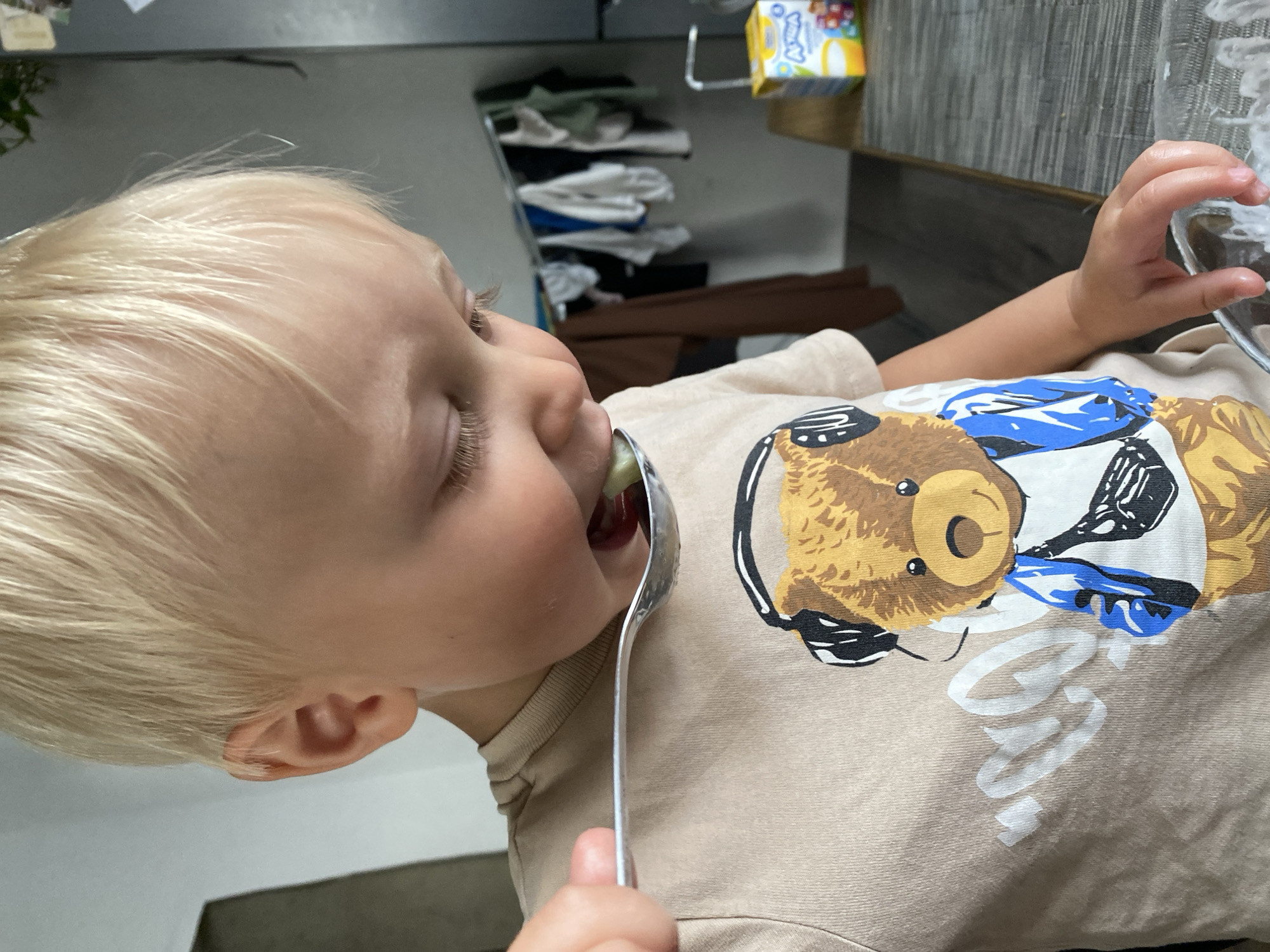 A close-up, eye-level shot shows a young Caucasian child with blonde hair, leaning forward as they bring a spoon of food to their mouth. The child is wearing a tan t-shirt with a graphic of a teddy bear wearing headphones. In the background, a clothes rack with various garments is visible, along with a box of what appears to be baby food. The child's left hand is also visible, holding the spoon. The scene appears to be indoors, likely in a kitchen or dining area, based on the presence of a table and what looks like a refrigerator. The child's eyes are looking down at the spoon, and their mouth is open.