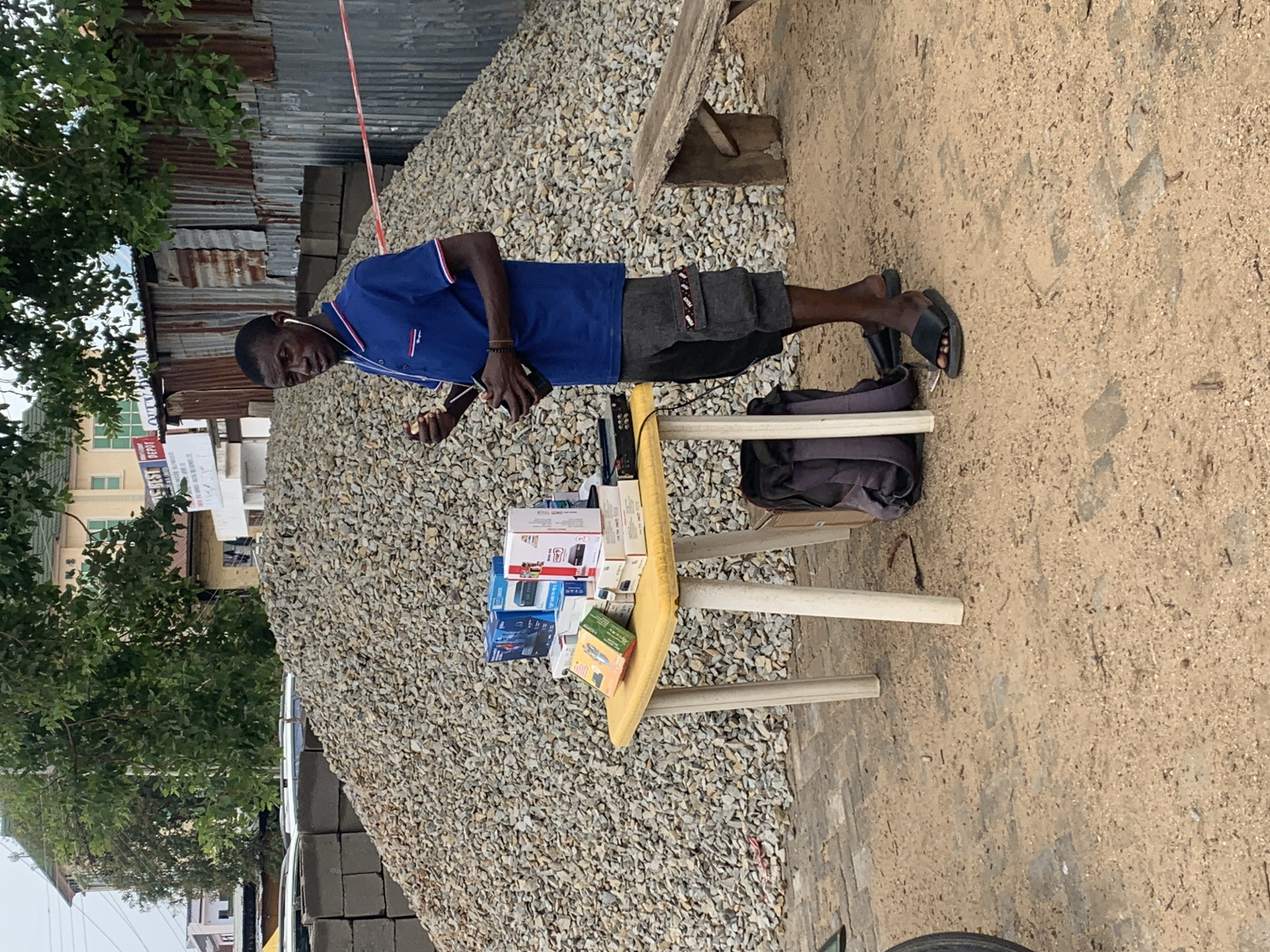 A man stands at a small table on a roadside in what appears to be Jos, Nigeria. He wears a blue collared shirt, dark shorts, and sandals, and has earphones in. On the table are several boxes of what appear to be electronics, along with a small radio or electronic device. Behind him is a large pile of gravel and corrugated metal fencing. A bag rests on the ground next to the table. The ground is a mix of dirt and small stones. Trees and buildings are visible in the background.