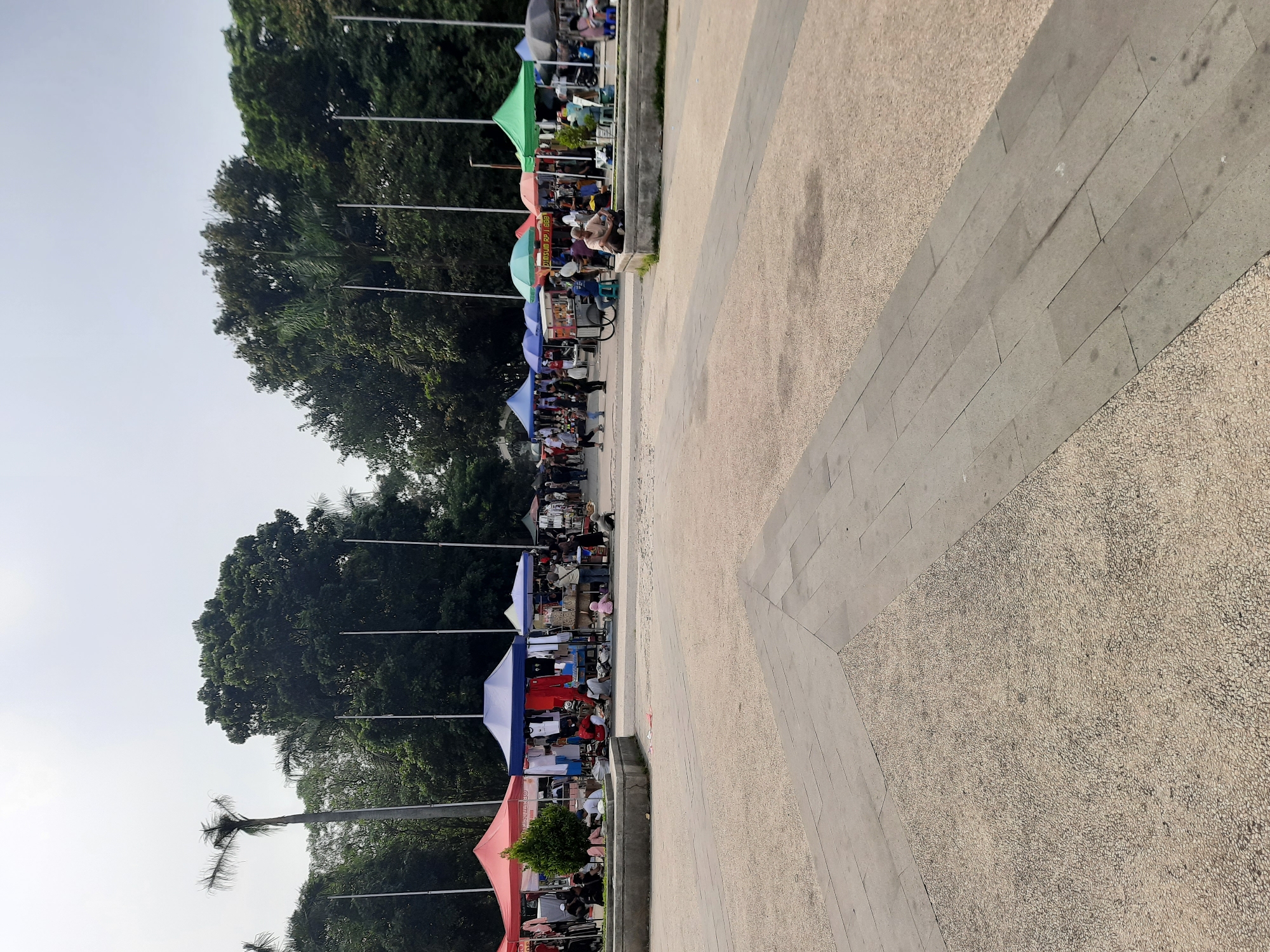 This is an outdoor market scene set in a paved public space in Bandung City, Indonesia. The market features numerous stalls with colorful tents, including red, blue, green, and orange. These stalls are displaying various goods, likely clothing and other retail items, with people browsing and interacting. Several individuals are seated or standing around the stalls. In the background, a dense line of trees forms a natural backdrop, with tall palm trees also visible. The ground is a mixture of gravel and concrete paving stones. The sky is overcast.