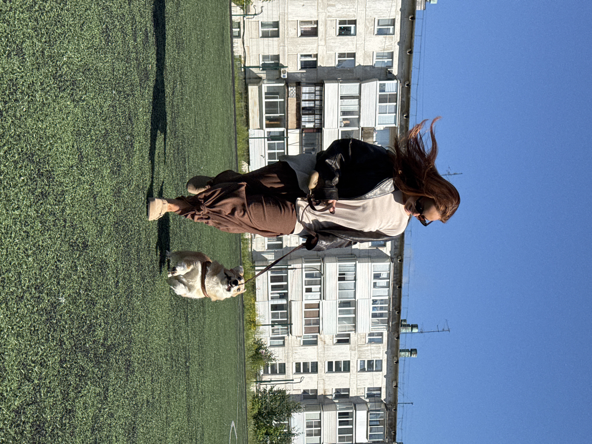 A woman with long, auburn hair, wearing sunglasses, a light-colored shirt, a black leather jacket, and brown pants is running on a green, artificial turf field. She is holding a leash attached to a small, tan and white corgi that is also running in front of her. Behind them, a tall, multi-story apartment building with a light-colored facade and numerous windows stands under a clear blue sky. The building appears to be a typical Soviet-era residential block, characteristic of cities like Chaikovsky, Russia. The field has markings for a sports pitch. The image captures a moment of outdoor activity on a sunny day.