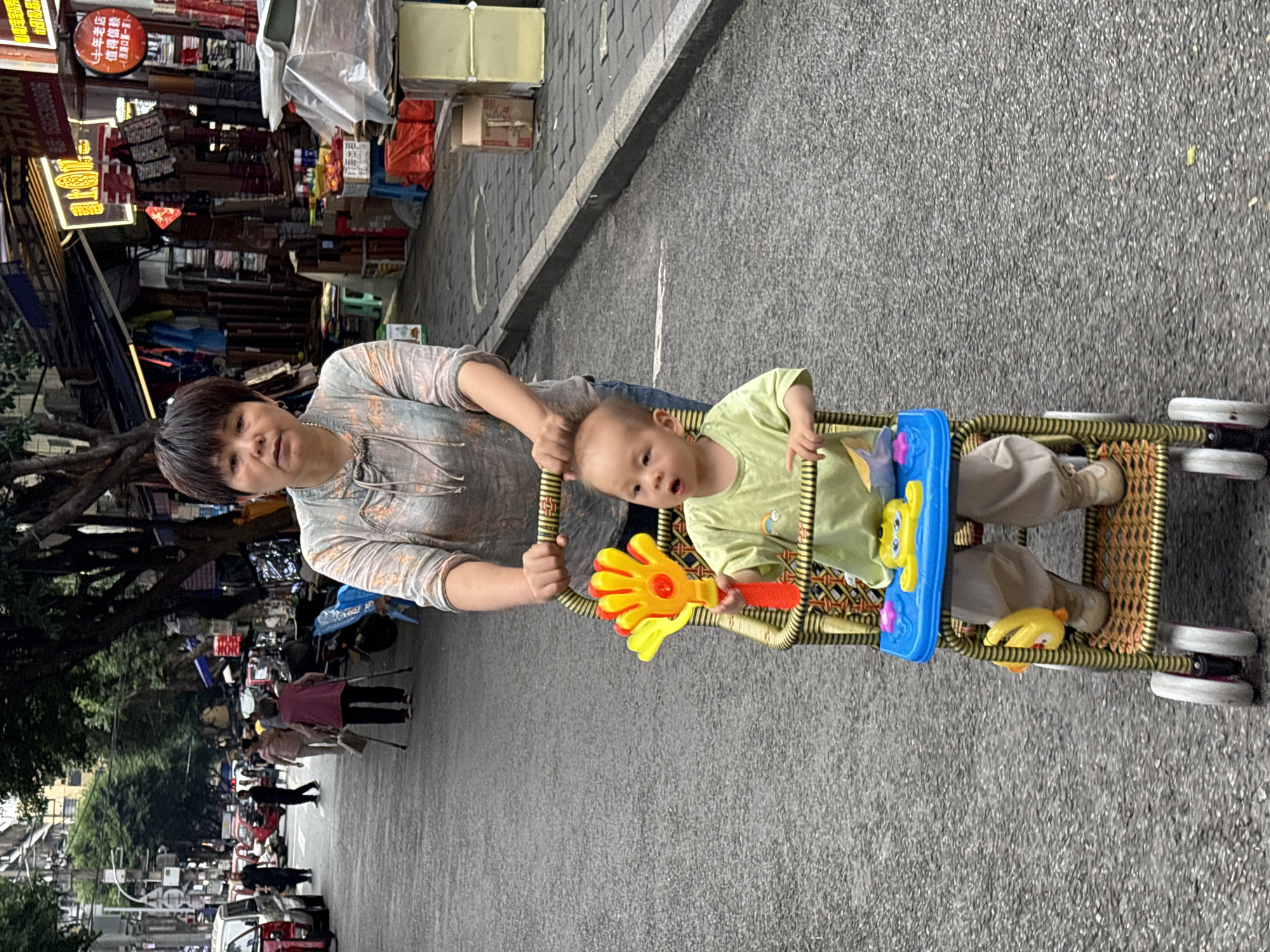 A Chinese woman, with short dark hair and wearing a grey patterned blouse, pushes a woven baby stroller down a paved street in Jiangjin, China. Inside the stroller, a young child in a light green t-shirt and beige pants looks forward with an open mouth, holding a yellow toy. The stroller has a blue tray with yellow plastic toys. In the background, other pedestrians walk on the street and sidewalks lined with shops. The street is an asphalt surface with visible tire marks. Buildings with awnings and signage are visible on the right side of the image. The overall atmosphere appears to be a bustling urban environment.