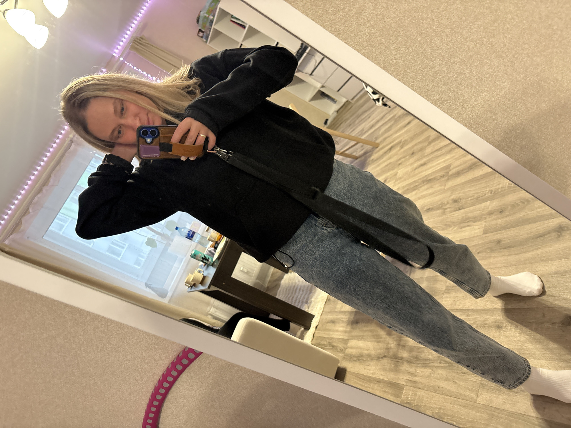 A woman is taking a mirror selfie indoors. She has blond hair, wears a black jacket, blue jeans, and white socks. She holds a smartphone in her right hand and rests her left hand on her face. The background includes a window, desk, shelving, and a strip of LED lighting. The location is indicated as Кондратово, Russia, although this is not discernible from the image content.