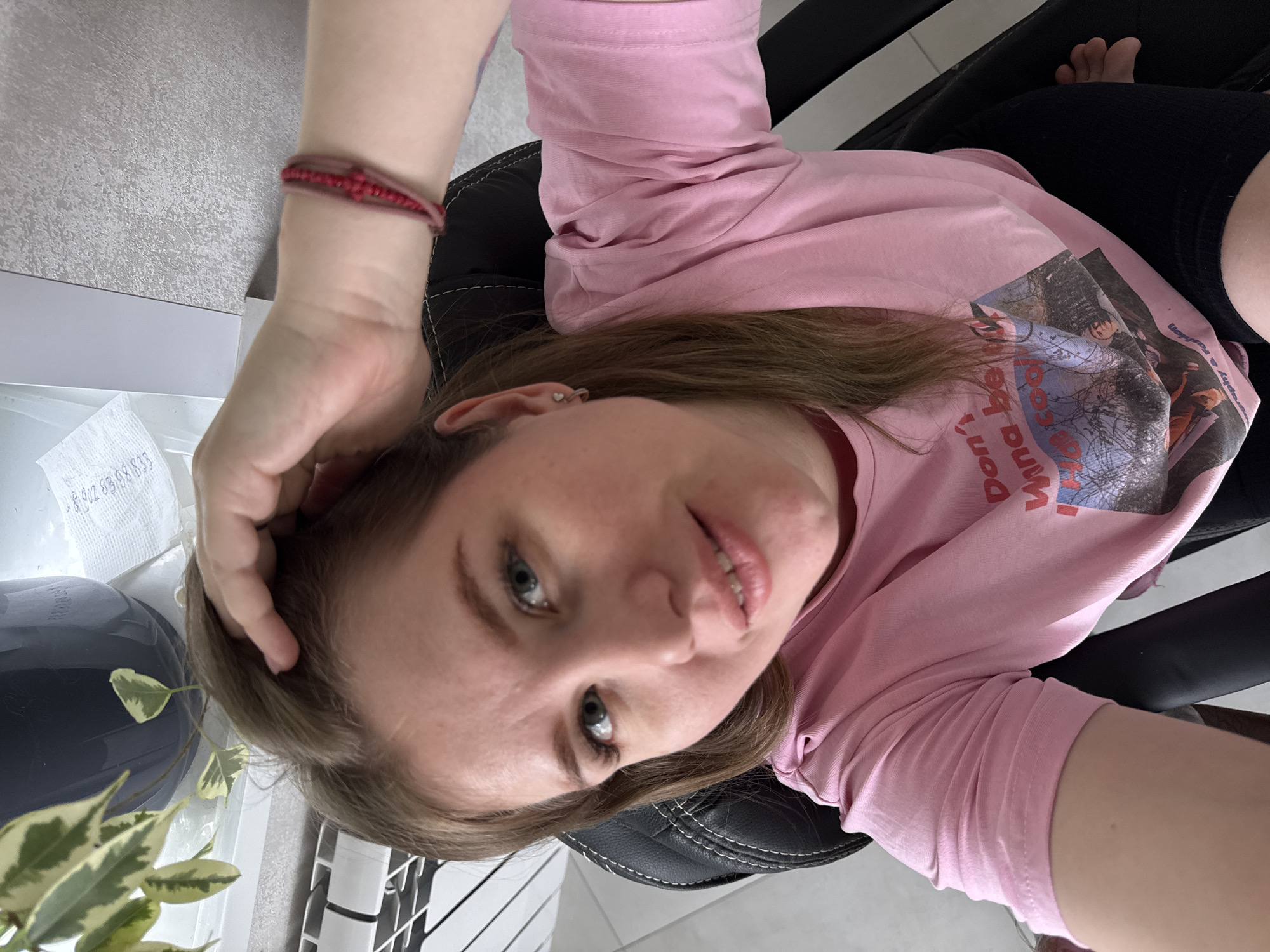 A woman is depicted indoors, seated in a black chair with white stitching, taking a selfie from an overhead angle in the city of Gornyi (Dvurechenskoe S/P), Russia. She has light-brown hair, light-colored eyes, and looks directly at the camera with a slight smile. Her attire consists of a pink short-sleeved t-shirt, partially displaying the text "Don't wanna be" and a multi-person graphic, and black legwear. A red beaded bracelet is on her right wrist, and a silver-colored earring is in her left ear. Her right hand is positioned with fingers touching her hair near the top of her head. The background includes a window sill in the upper left, supporting a dark-potted plant with variegated leaves and a crumpled white paper. A white heating radiator is partially visible below the window sill. The wall to the upper right is light grey with a textured finish. The floor, visible in the lower right, is light-colored tile.