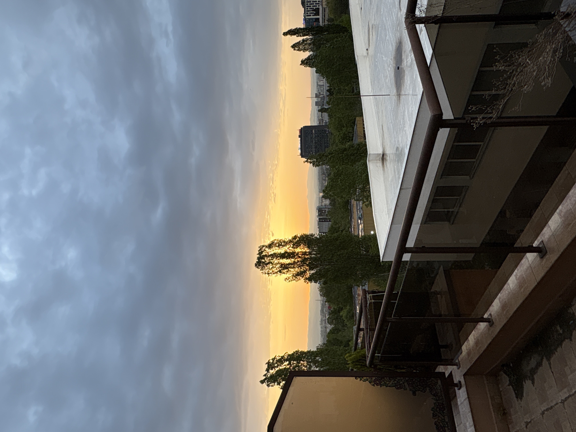 The image depicts a view of Sofia, Bulgaria at sunset from a balcony. The sky is filled with dark clouds on the left, transitioning to a bright, golden hue near the horizon where the sun is setting. Silhouetted against the sunset are trees and buildings of varying heights. In the foreground, a portion of a building with a white flat roof and a metal railing balcony is visible. The balcony is attached to a building with stone walls and a tan facade. The scene is still and quiet.