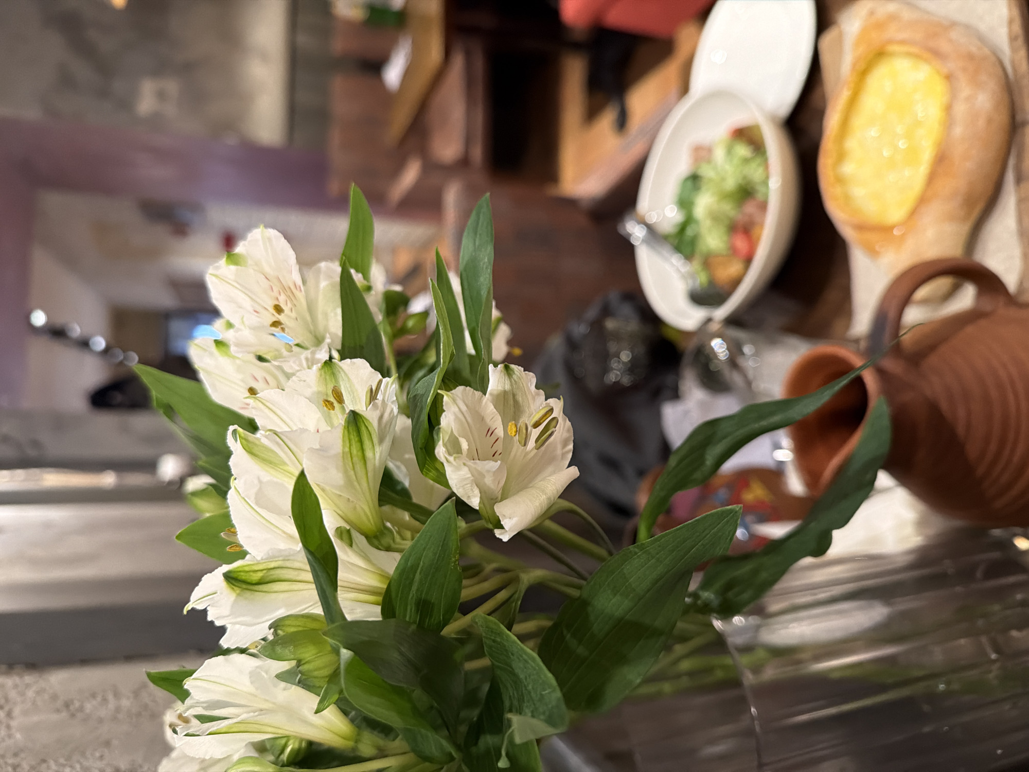 The image presents a close-up view of a bouquet of white Alstroemeria flowers with green leaves, occupying the left and central foreground. Behind the flowers and to the right are indistinct food items, including what appears to be a bread boat filled with yellow cheese, next to a white oblong bowl containing a salad. Adjacent to these food items is a brown clay pot. The flowers are in a clear vase, set on a dark surface. The background consists of blurred interior elements, which suggest a restaurant or dining area. The location, St. Petersburg, Russia, is not discernible from the visual information available in the image. There are no people visible. The interactions appear to be an arrangement of objects on a surface, likely in preparation for a meal. The environment suggests an indoor setting with controlled lighting.