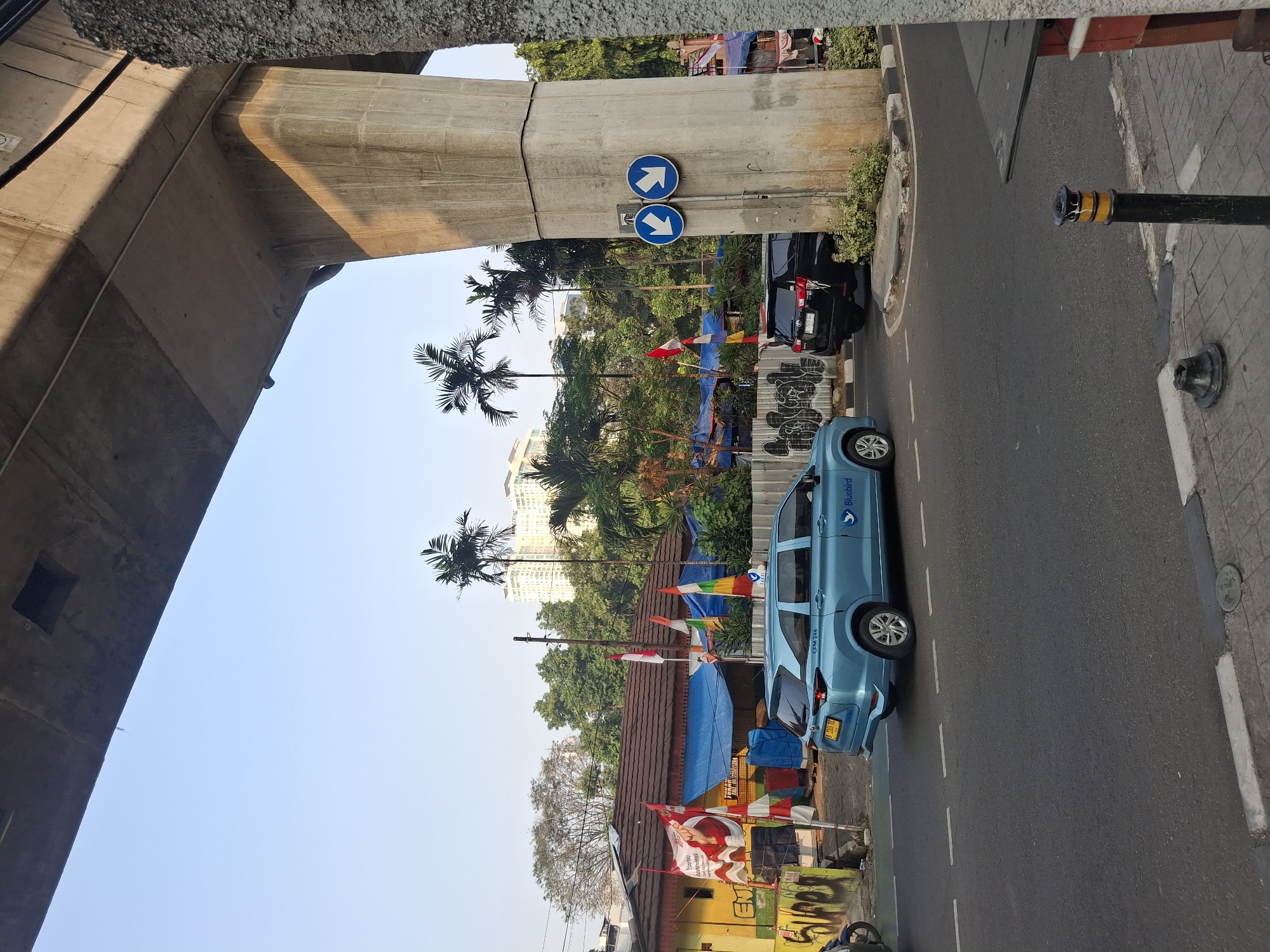 A light blue Bluebird taxi is seen driving on a paved road in South Jakarta, Indonesia, under the shadow of an elevated concrete structure. To the right of the taxi, a black SUV is also present. Two blue circular traffic signs with white arrows indicating a two-way traffic direction are mounted on a concrete pillar supporting the elevated structure. In the background, there are buildings, trees, and flags, including Indonesian flags. The scene appears to be an urban environment with clear skies.