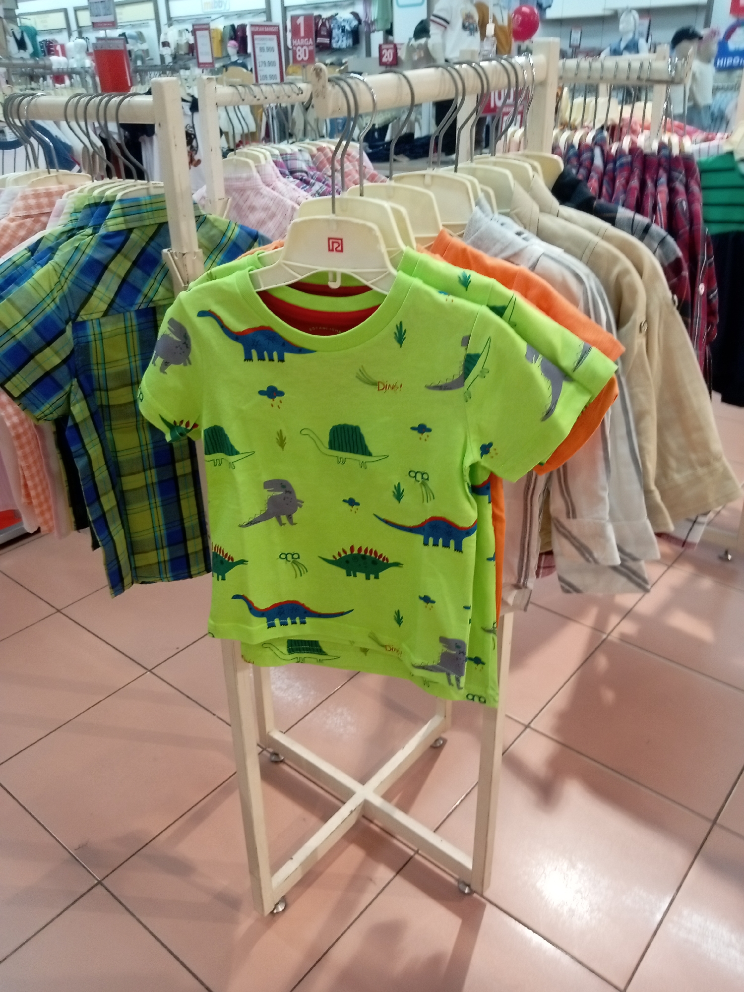 The image shows a display of children's clothing in what appears to be a retail store in Batam, Indonesia. In the foreground, a bright lime green t-shirt with a dinosaur print is prominently displayed on a white hanger. The t-shirt features various illustrations of dinosaurs in blue, grey, and green. Behind this t-shirt, other clothing items are visible on hangers, including a plaid shirt in green and blue, and other shirts in various colors and patterns. The clothing is hung on a multi-tiered rack, with a white metal frame structure supporting the hangers. The background reveals more clothing racks, display shelves with merchandise, and store signage. The floor is tiled with large, light pinkish-beige square tiles. The lighting suggests an indoor environment, typical of a retail store. No people are visible in the frame. The overall scene depicts a merchandising display in a clothing store, likely for children.