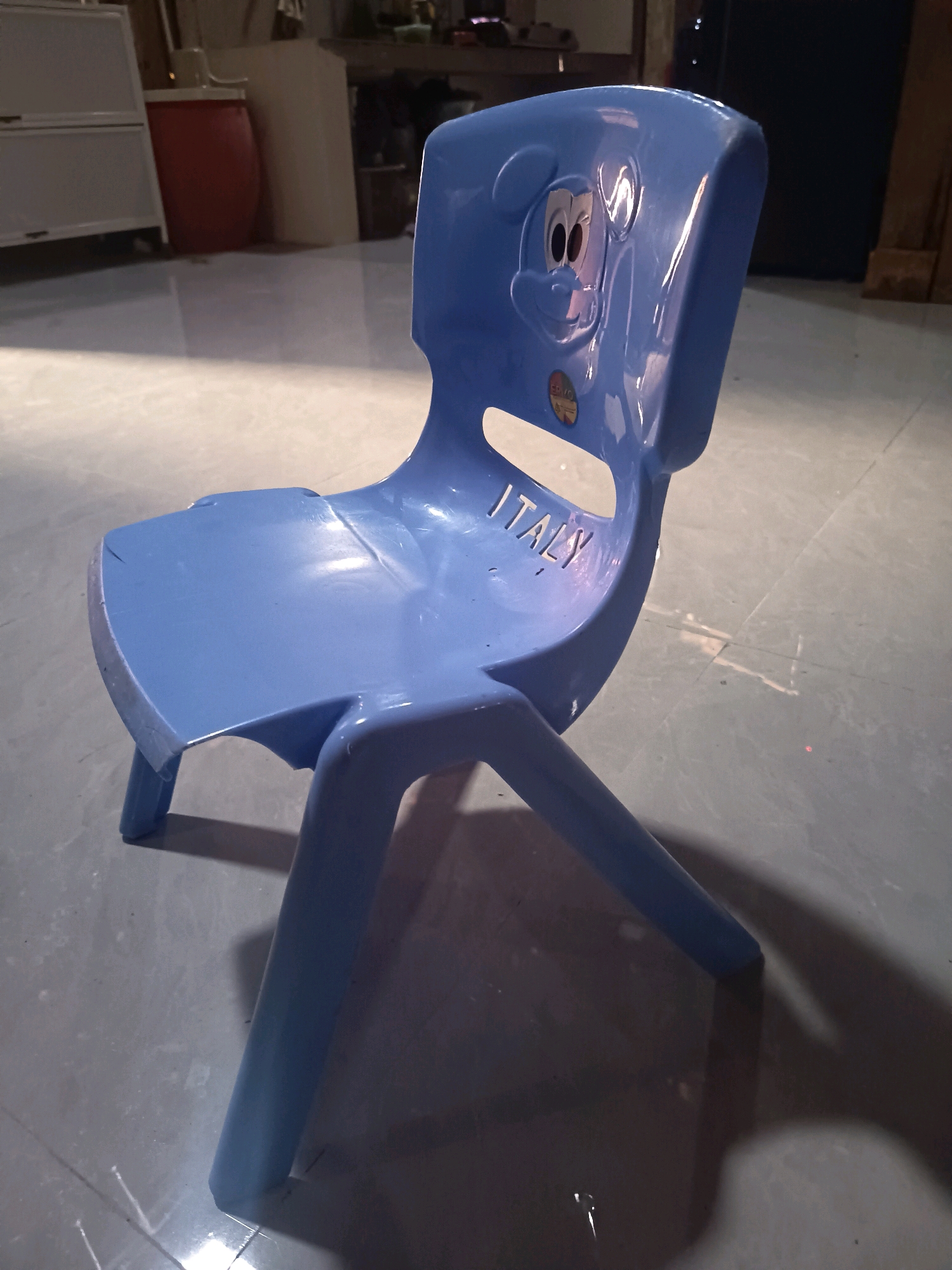 A light blue plastic chair with a Mickey Mouse design on the backrest and the word "ITALY" cut out of the backrest is positioned in the center of the frame. The chair is on a tiled floor that reflects the light. In the background, there are glimpses of shelves, a red barrel, and a dark wall, suggesting an indoor or covered outdoor setting. The lighting is somewhat dim, with strong highlights and shadows on the chair and floor. The overall scene is still and devoid of any human activity. The location is not specified in the image.