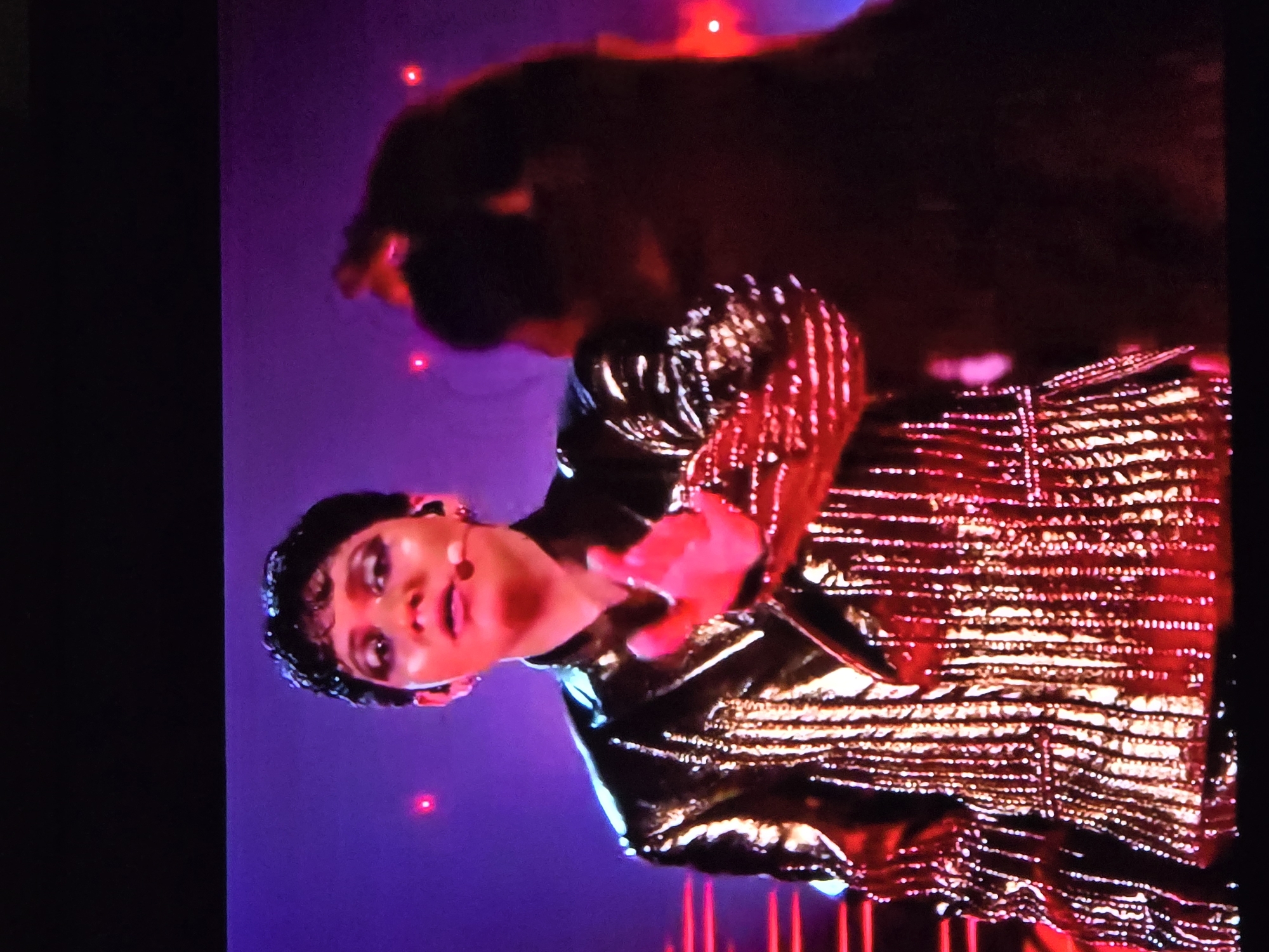 A person with short, dark hair is wearing a gold, metallic jacket and appears to be singing or speaking into a microphone. The person's skin tone is light. The background is purple with red lights. Another person with dark hair is standing behind the first person. The location is unknown.