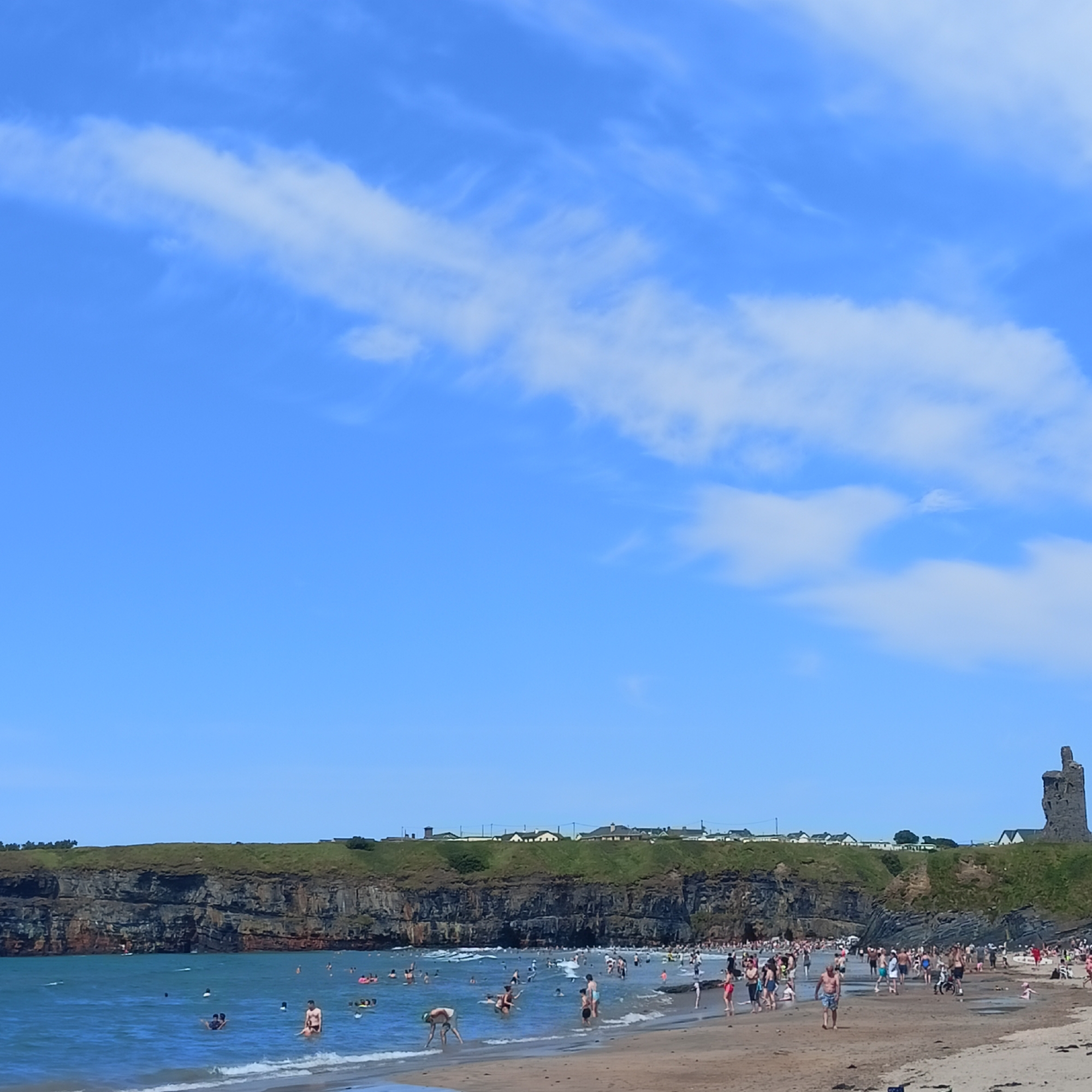 The image depicts a crowded summer day at Ballybunnion Beach in Ballybunnion, Ireland. A wide sandy beach is filled with numerous people of all ages. Some are sunbathing, while many are in the water, swimming, playing, or wading. The sea is a vibrant blue, with small waves lapping at the shore.
Behind the beach, a dramatic cliff face rises, covered in green grass and dotted with buildings along its edge. The cliff is a prominent feature of the landscape. To the far right of the image, a distinctive, weathered stone tower stands on top of the cliff, a historical landmark of the area. The sky is a clear, bright blue with scattered white clouds. The overall scene is one of a lively and popular tourist destination enjoying good weather.