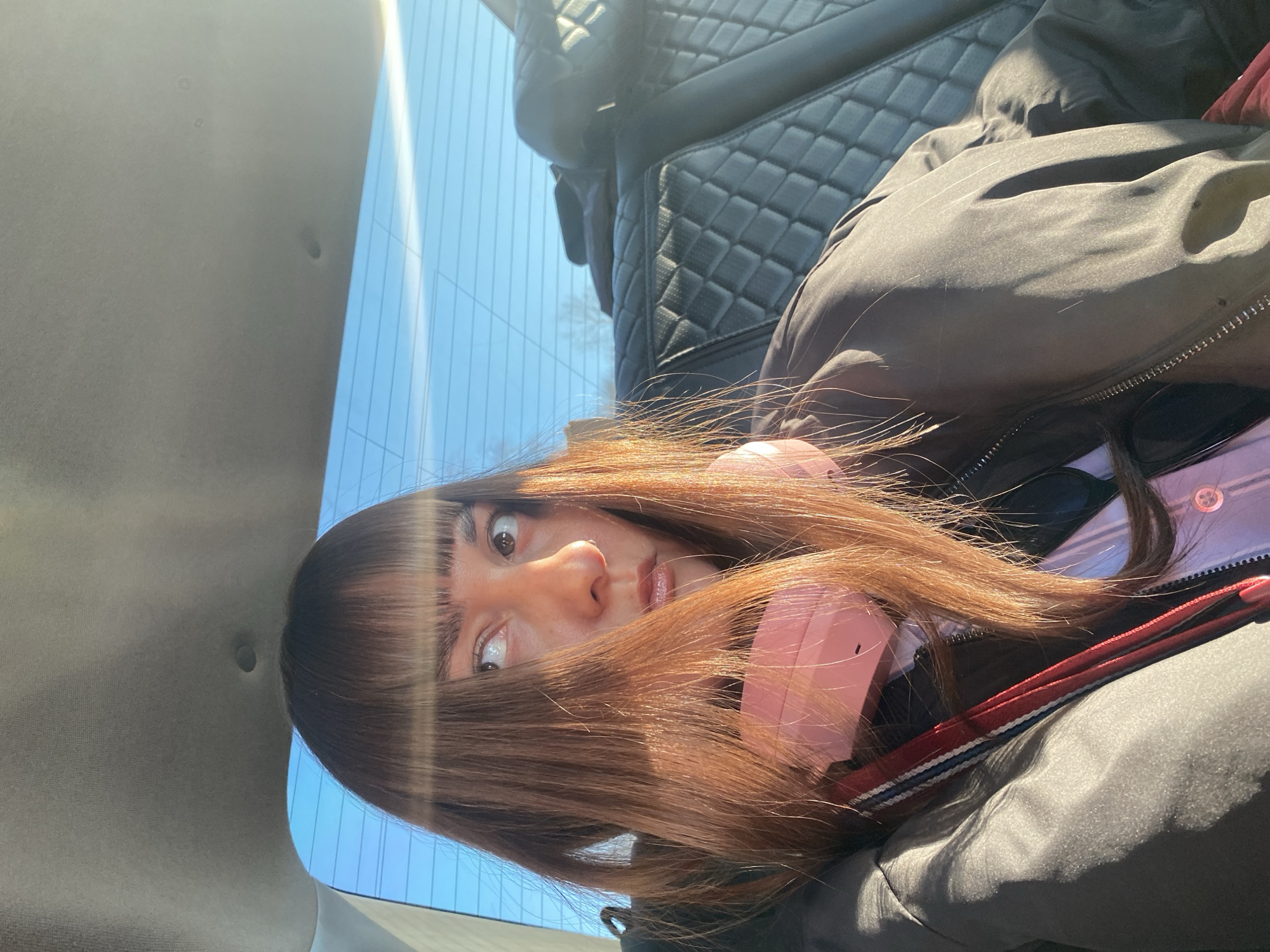 A person with brown hair, wearing pink headphones, is sitting in a car. The car is parked beside a building in Votkinsk, Russia, seen through the window. The interior of the car features black leather seating with a quilted pattern. The person is wearing a dark jacket over a pink shirt.