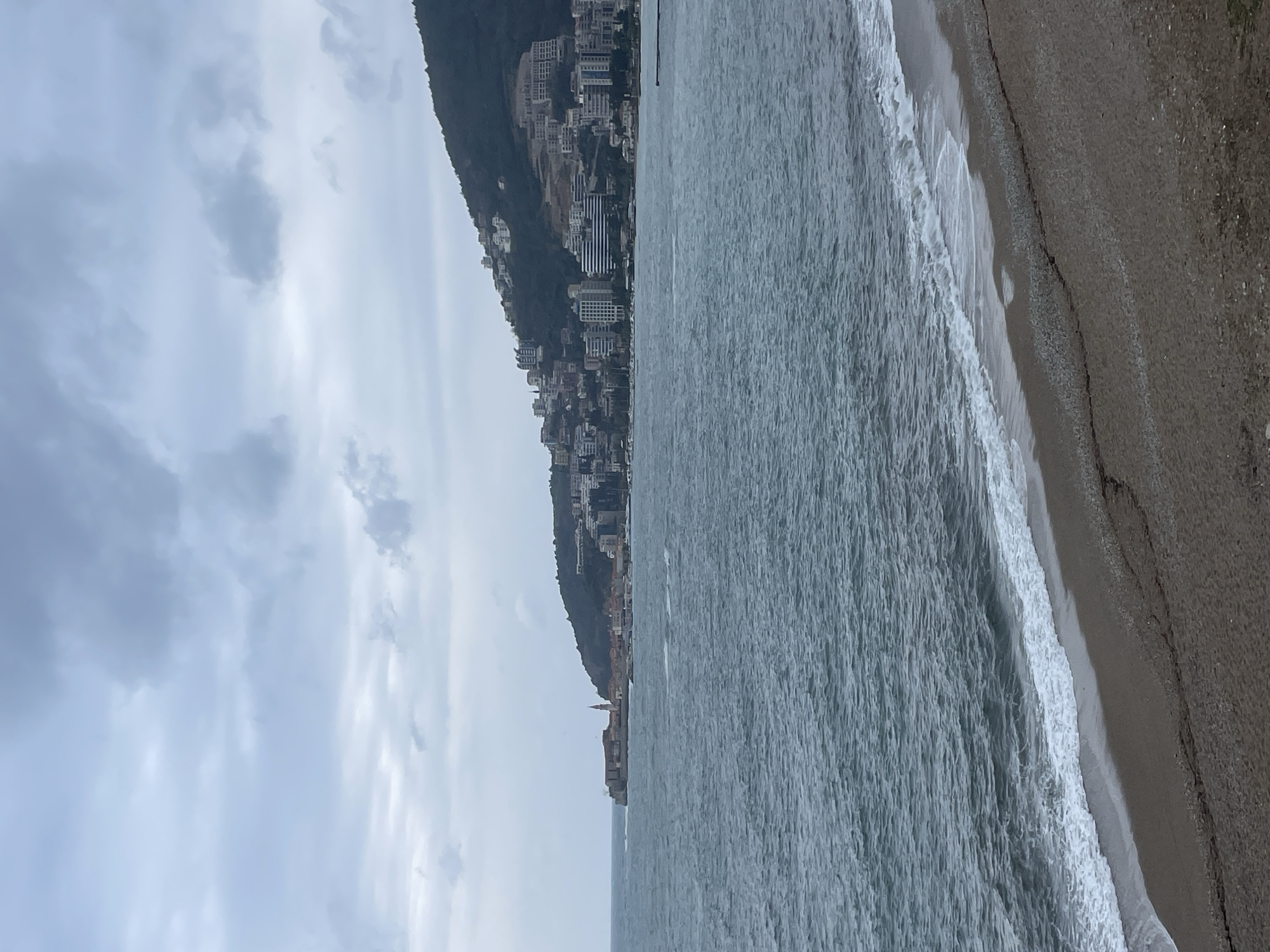 The image depicts a panoramic view of the coastline and cityscape of Budva, Montenegro, observed from a sandy beach under an overcast sky. The foreground features a dark brown-grey beach with fine pebbles, where light blue-grey waves of the Adriatic Sea break, forming a line of white foam. The midground is dominated by the expansive, turbulent sea. To the left, a fortified peninsula, identifiable as Budva's Old Town (Stari Grad), extends into the water, featuring historical buildings and a prominent bell tower. To the right, a large, dark green hill slopes upward, densely populated with numerous multi-story modern residential and commercial buildings. Several construction sites with exposed earth are visible on the hillside. The sky is uniformly light grey, filled with low, diffused clouds, indicating an overcast condition. No individuals or specific vessels are discernible within the frame.