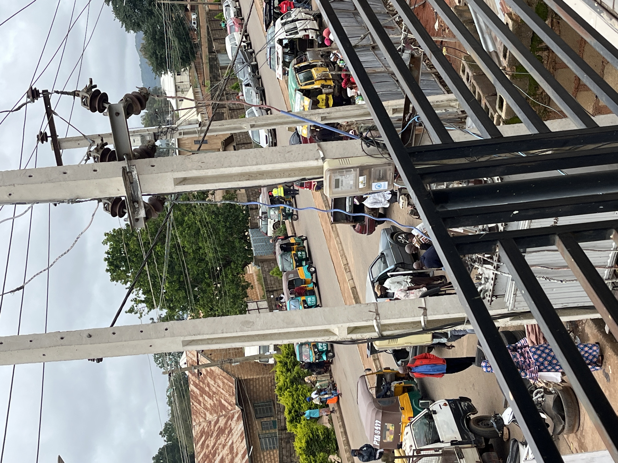 The image depicts a street scene in Bukuru, Nigeria, on an overcast day. The foreground is dominated by a black metal railing, suggesting the vantage point is from a balcony or elevated structure.
In the midground, a prominent concrete utility pole stands vertically, adorned with multiple insulators and electrical wires crisscrossing the sky. A meter box is attached to the pole. The street is lined with various vehicles, including several three-wheeled auto-rickshaws, commonly known as "Keke Napep," some of which are yellow and green, while others are brown. One brown auto-rickshaw has the license plate "PTA-8477". There are also cars and a white pickup truck visible.
People are present on the street and sidewalk. Some are standing near the auto-rickshaws, possibly as passengers or drivers, while others appear to be walking. A woman in a blue and pink patterned dress walks with a white bag. A man in a red jacket and blue shirt is standing on the road.
The background reveals buildings with corrugated iron roofs, some appearing aged. Lush green trees and bushes are interspersed throughout the scene. In the far distance, hills are visible beneath the cloudy sky. The overall impression is of a busy urban environment with active street life and a somewhat weathered aesthetic.