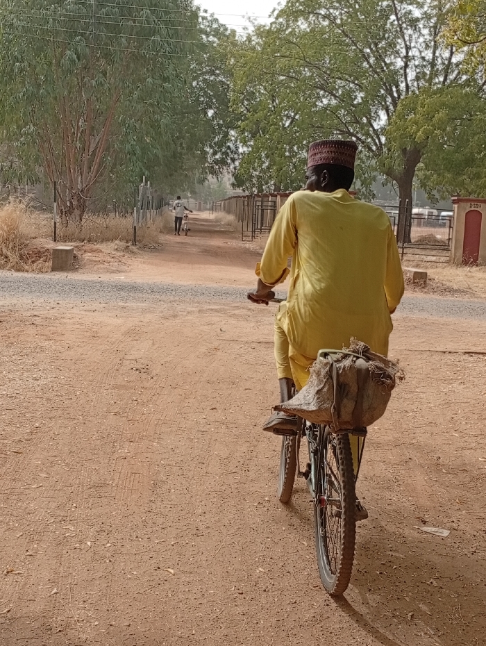 From the back, a man in a yellow robe and a reddish-brown cap is riding a bicycle down a dirt path. The bicycle has a large sack or bag tied to the rear carrier. In the distance, another person is walking and pushing a smaller bicycle in the same direction. Trees line both sides of the path and the sky is lightly clouded. A building is visible on the right of the path near the rider.