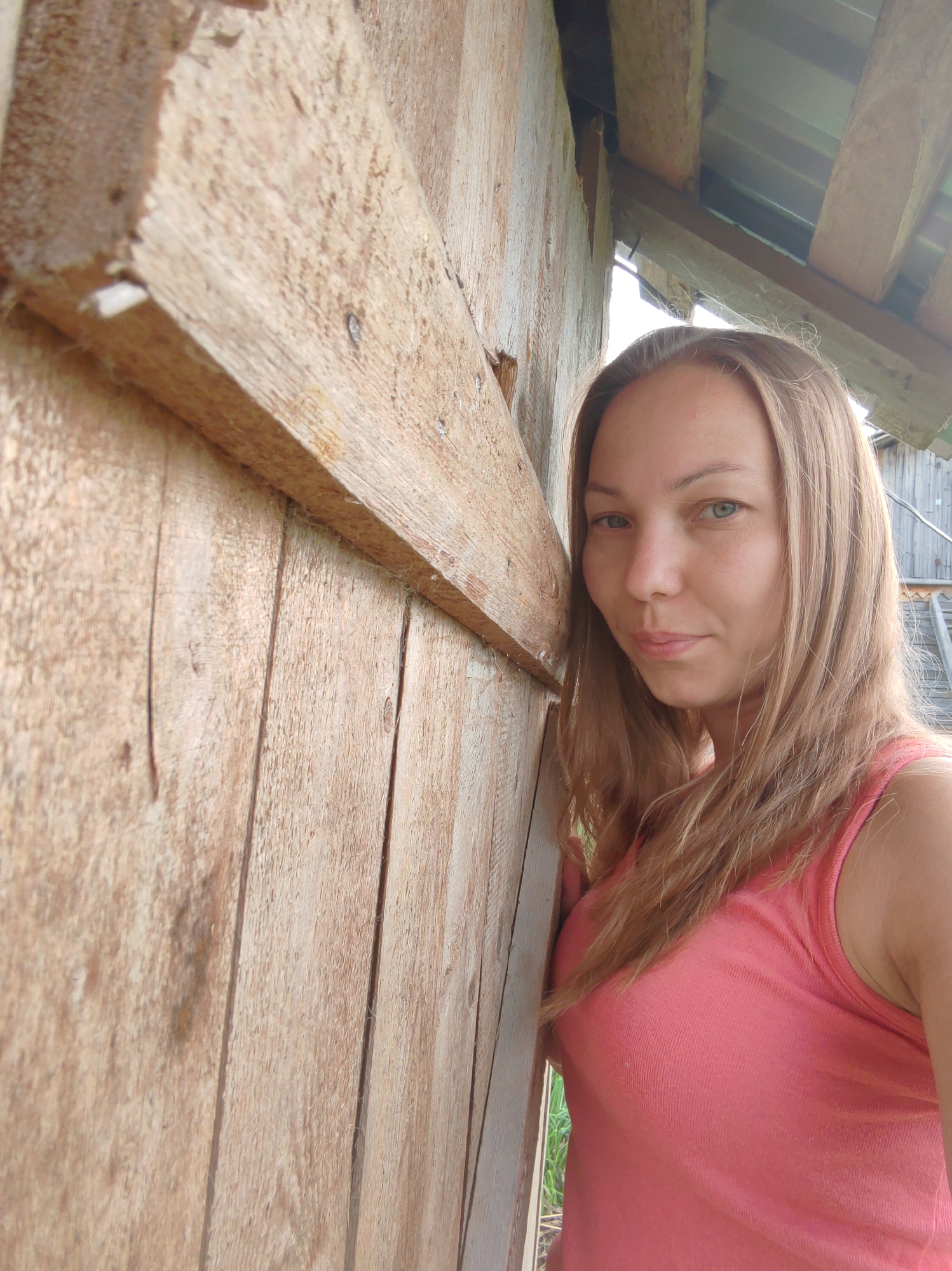 A blonde woman in a coral tank top leans against a weathered wooden structure. The background shows more wooden beams and what appears to be a corrugated metal roof, suggesting an outdoor, possibly rural or agricultural setting. The image is a close-up, with the woman's face and upper body prominently featured. The wooden planks of the structure are rough and show signs of age. The lighting suggests daylight.