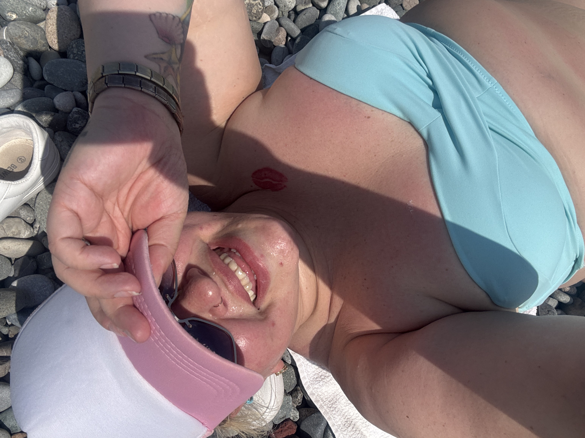 In a close-up, top-down shot, a person wearing a light blue bikini top and a white and pink baseball cap basks in the sun on a pebbled beach. Their hand is raised to the brim of the cap, partially obscuring their face, which is turned upwards towards the camera, revealing a wide smile and sunglasses. A faint red lipstick kiss mark is visible on their chest. The background consists of smooth, gray and dark pebbles. A white shoe is visible in the upper left corner of the image. The scene suggests a relaxed day at the beach in Batumi, Georgia.