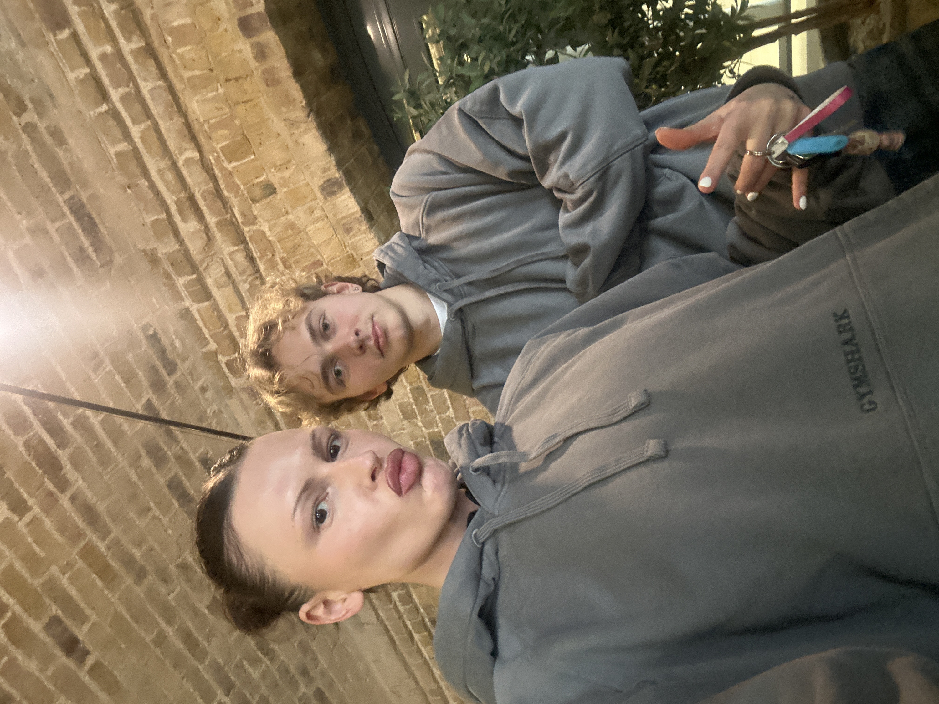 An indoor scene depicts two individuals, an adult female and an adult male, positioned centrally against a background of an arched brick ceiling, characteristic of converted railway arches or similar structures in London, United Kingdom.
The female, positioned on the left and closer to the camera, has fair skin, dark hair pulled into a bun, and is making a "kissy face" by pursing her lips. She wears a grey hooded sweatshirt with "GYMSHARK" printed in dark grey on the chest. Her right hand is visible in the lower right, displaying white-painted fingernails and holding a set of keys attached to a metallic ring, a blue fabric fob, a pink and white striped lanyard, and a small, light-colored charm.
The male, positioned slightly behind and to the right of the female, has fair skin, curly blonde hair, and a neutral expression, looking directly at the camera. He wears a plain grey hooded sweatshirt over a white t-shirt, with his arms crossed.
The overhead environment features light brown brickwork laid in an arching pattern, with distinct mortar lines. A vertical, dark metal fixture or cable extends from the ceiling on the left. Artificial light illuminates the scene from above. Behind the male subject, a dark, possibly reflective, horizontal surface is visible, beneath which green foliage from potted plants is present.