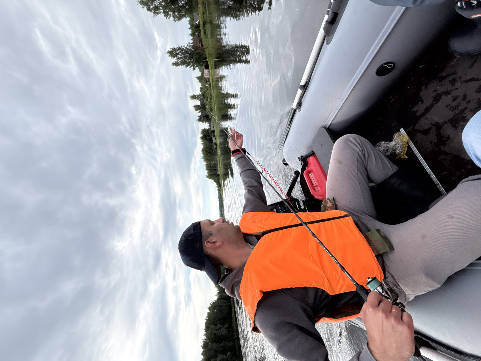 A man in a grey hoodie and an orange life vest sits in a grey inflatable boat, fishing on a calm river. He wears a black baseball cap and is holding a fishing rod. The boat is moving, creating ripples on the water. In the background, the riverbank is lined with trees and houses, suggesting a rural or semi-rural setting. The sky is cloudy and overcast. A red fuel can is visible in the boat. The scene appears to be located in or near Anteplevo (Okhotinskoe S/P), Russia.