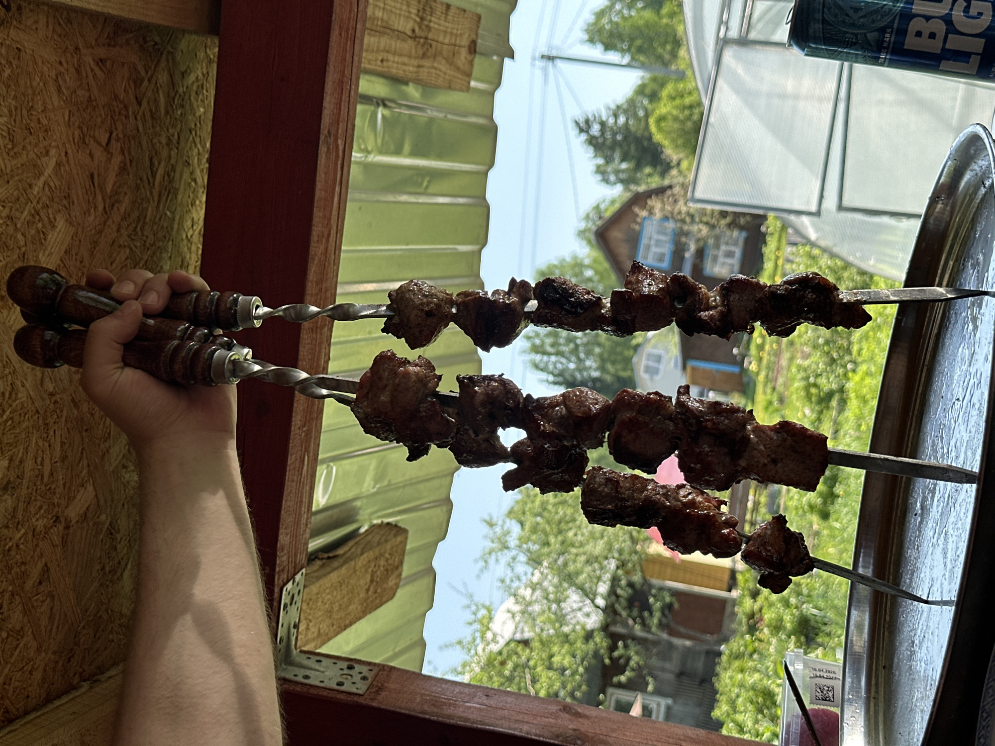 A man is holding two skewers of cooked meat in front of a partially constructed structure with a view of a residential area in Khokhryaki, Russia. The skewers have brown, carved wooden handles and twisted metal rods with chunks of dark, grilled meat. The man's forearm is visible, light-skinned and slightly hairy. The structure appears to be under construction, with corrugated metal sheets, wooden beams, and red-painted wooden posts. Beyond the structure, there is a view of houses with blue window frames, trees, and a blue sky. A can of Bud Light and a metal tray can be seen to the right. A small plastic container with writing is also visible.