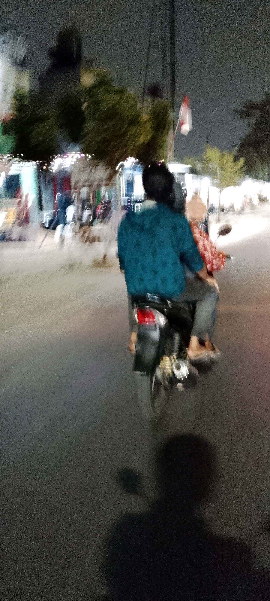 The image shows two individuals riding a motorcycle at night on a street in the city of Bekasi, Indonesia. The person in the front, who appears to be driving, is wearing a teal-colored shirt and a helmet. The person in the back is wearing a red patterned shirt and has their arm around the driver. The motorcycle's taillight is visible, as are its wheels and exhaust pipe. In the background, there are trees, streetlights, and buildings, suggesting an urban or semi-urban environment. The lighting indicates it is nighttime, with artificial lights illuminating the scene. A shadow of the motorcycle and its riders is cast on the road in the foreground. The overall image has a grainy texture due to the low-light conditions.