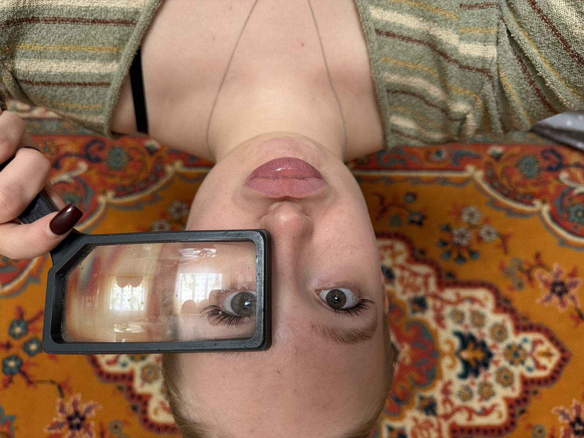 An overhead, inverted close-up shot features a person, likely female, lying on a vibrant, patterned rug in an indoor setting in Засечное, Russia. The individual's head is towards the bottom of the frame, with their neck and chest extending upwards. Their left hand, adorned with dark red nail polish, holds a black-framed magnifying glass over their left eye.
The person has fair skin, a neutral facial expression, and plump lips with a glossy, light pink lipstick. A delicate silver necklace rests on their chest. Their eyes are light-colored (grey-green), and long, dark eyelashes are prominent, especially magnified through the lens on the left eye. The reflection in the magnifying glass shows clear window frames, indicating daylight from an external source.
The individual wears a loose-fitting, striped top or cardigan in shades of sage green, beige, and reddish-brown. The background and surface on which the person is lying is a richly patterned rug, featuring intricate floral and geometric motifs in warm tones of orange, red, blue, and cream. No visible text is present in the image. The scene suggests a moment of playful curiosity or close examination.