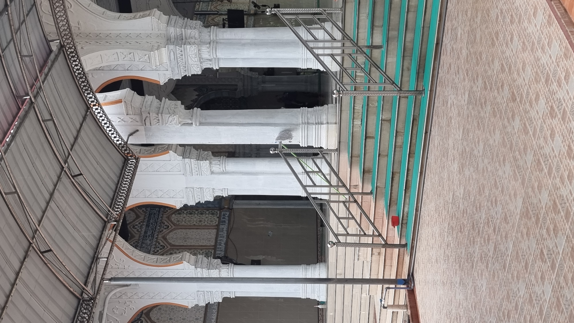 The image shows the entrance to a building with white pillars and arched openings, likely a mosque in Meunasahlhok, Indonesia. A metal roof supported by a framework covers the area above the entrance. In front of the building, a set of stairs leads up, with stainless steel railings on either side. The stairs are covered with green mats. The ground in the foreground is tiled. A red bucket sits on one of the steps. The overall impression is of a clean and well-maintained public space.