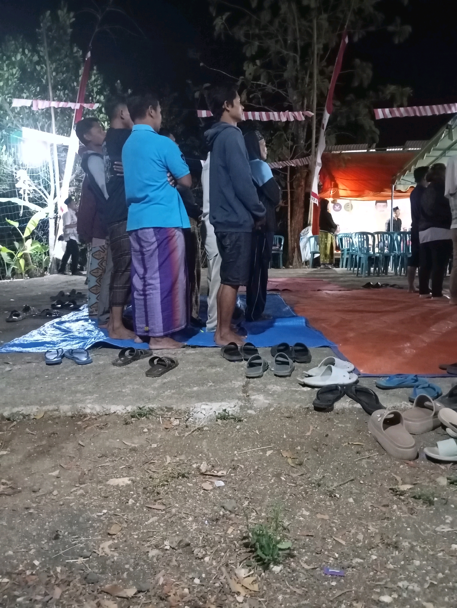 In Wonogondo, Indonesia, a group of people, mostly men and boys, are gathered outdoors at night. They are standing on a blue tarp laid on a concrete surface, with a larger orange tarp and a canopy in the background, suggesting an event or gathering. Several individuals are wearing sarongs, traditional Indonesian lower garments, and others are in casual clothing. A variety of sandals and flip-flops are scattered on the ground in front of the group, indicating that they may have removed their footwear. The scene is illuminated by artificial lights, and trees with decorative red and white banners are visible. In the background, there are rows of blue chairs under the canopy, and a few people are visible near them, along with what appears to be a banner with a portrait. The ground in the foreground is covered with dirt, leaves, and small plants.