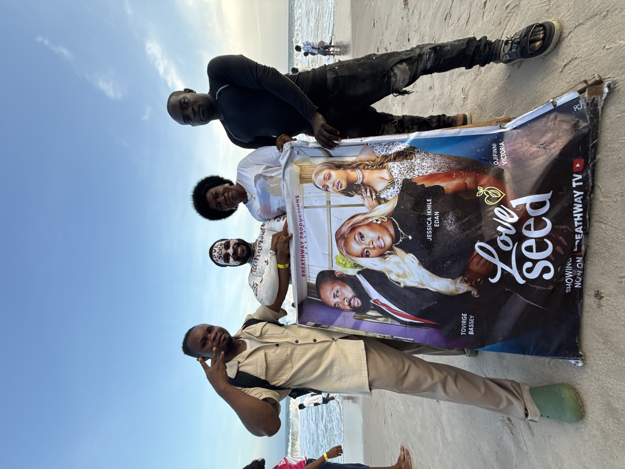 Four individuals stand on a sandy beach in Mopo Onijebu, Nigeria, holding a promotional poster for the film "Love Seed." The poster features images of actors Jessica Ikhile Edan, Tovirge Bassey, and Ojifinni Victoria. The man at the bottom of the frame is wearing a tan shirt and khaki pants, making a peace sign with his hand. The second man wears a patterned white shirt and face paint. The third person is a woman wearing a white top. The fourth man, at the top, is dressed in black. In the background, the sea meets the sky under a bright daylight. Other people are visible further down the beach.