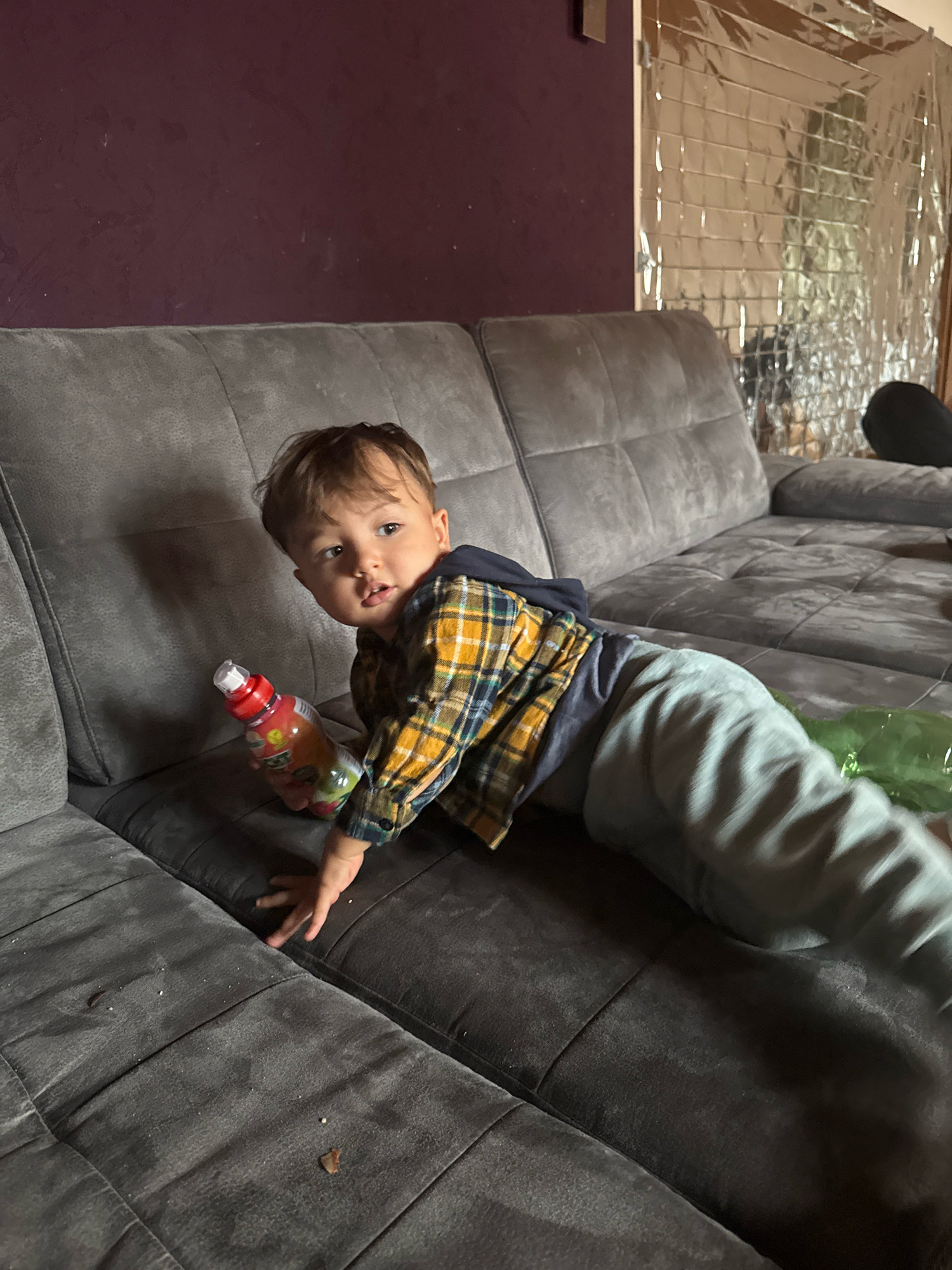 A toddler wearing a plaid shirt and grey pants is lying on a dark grey sectional couch, holding a red bottle with a white cap. The couch is positioned in a room with a dark purple wall on the left and a wall covered in reflective material with a grid pattern on the right. The toddler's body is stretched out along the length of the couch, and their head is turned towards the camera. A green object is visible on the couch to the right of the toddler. The floor is not visible. The scene appears to be indoors.