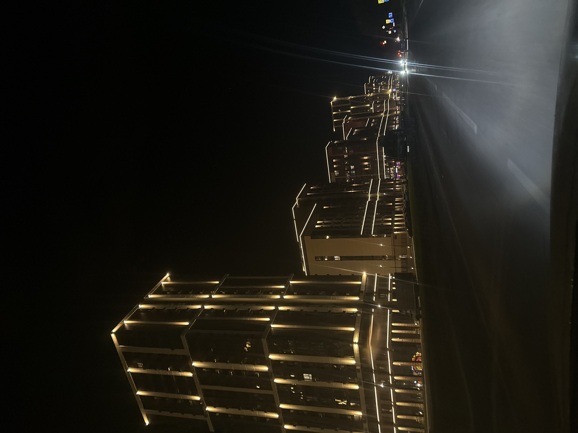 At night in Rostov-on-Don, Russia, a series of modern, multi-story buildings are illuminated with linear lights accentuating their architectural features. The buildings appear to line a roadway, which is also lit, exhibiting street markings. The sky is dark, suggesting a nighttime setting. The buildings are the primary focus, dominating the foreground and midground. Distant streetlights are visible further down the road.